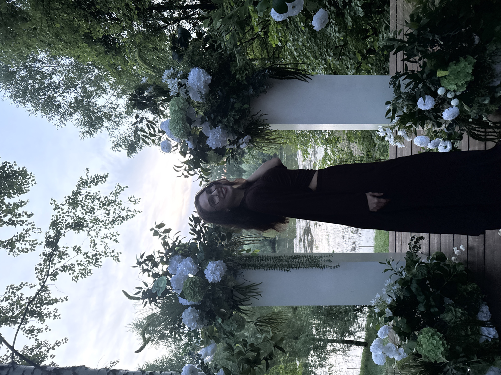 The image depicts a woman with long brown hair, wearing glasses and a dark dress, standing in front of two white pillars decorated with white and green floral arrangements. The pillars appear to be part of an outdoor structure, possibly for a wedding or event. Behind the pillars is a body of water, with trees and greenery visible on the opposite bank. The overall setting is outdoors, with foliage suggesting a park or garden environment. Based on the provided location, Эстачи, Russia, this scene is likely situated in that city. The woman is posing and smiling towards the camera.
