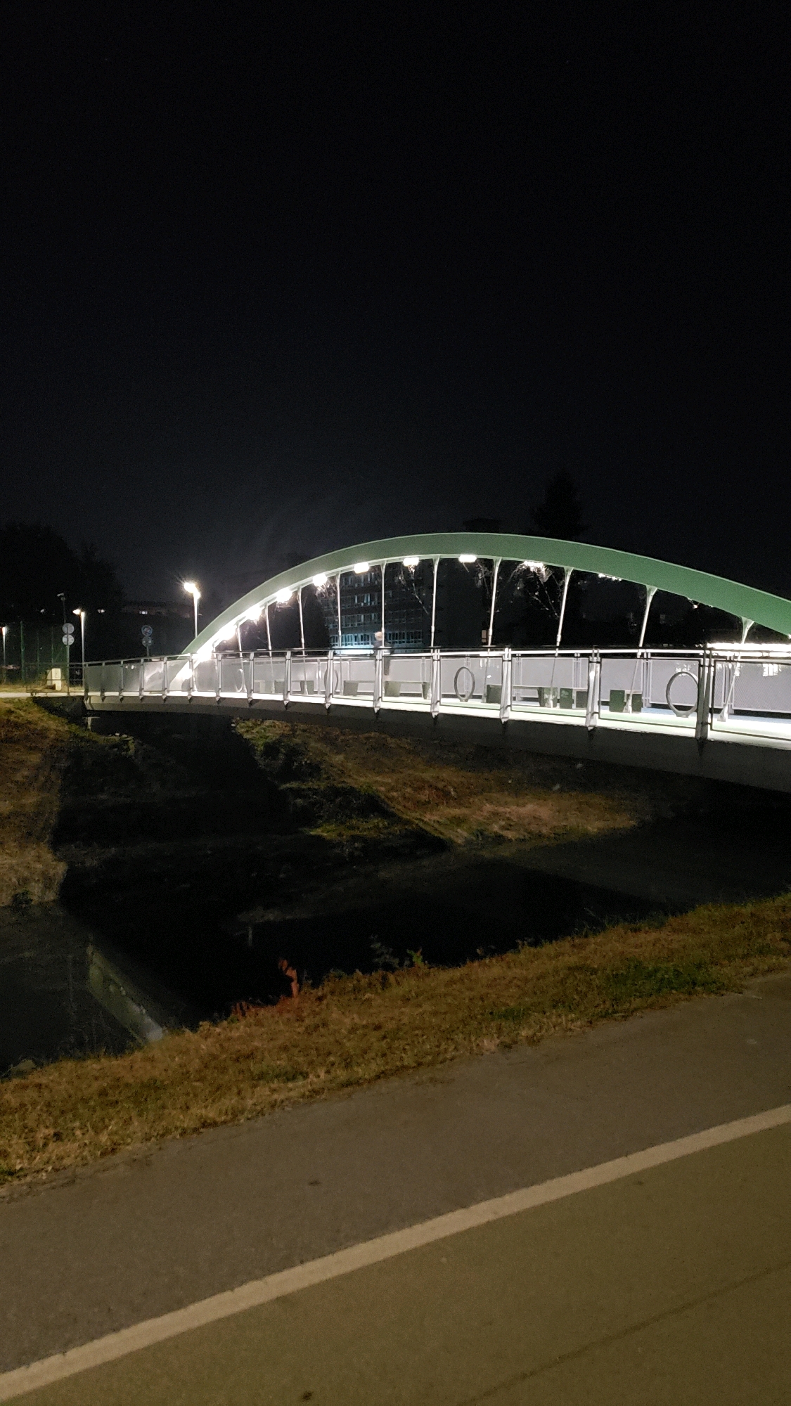The image shows a modern pedestrian bridge illuminated at night. The bridge has a green, arched canopy supported by white pillars and features a white railing with circular decorative elements. It spans across a dark body of water, likely a river or canal, with grassy banks. A paved pathway runs along the foreground of the image, adjacent to the grassy bank. In the background, a building with illuminated windows is visible. The overall scene is dark, with the bridge and building being the primary sources of light. This bridge is the pedestrian bridge over the Torysa River in Prešov, Slovakia.