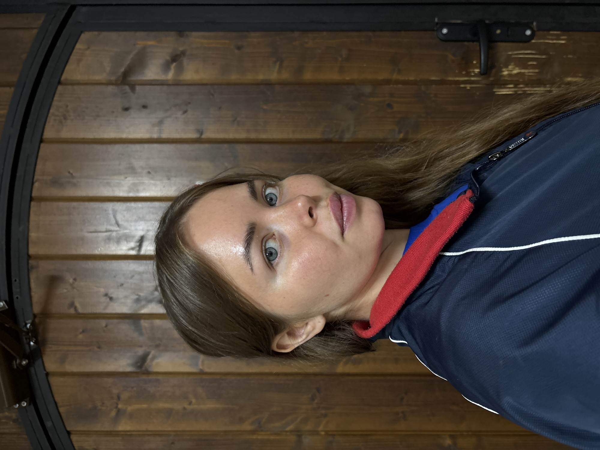 A woman with fair skin, blue eyes, and light brown hair is the central figure in the image. She is wearing a navy blue jacket with a red and blue collar and a white stripe. Her face is directed towards the camera, and she appears to be looking directly at the viewer.
The background consists of a wooden wall constructed from horizontal planks. A black metal frame is visible on the left side of the image, partially obscuring the wall. A black metal handle is present in the upper right corner.
The location is Русский Вожой, Russia.