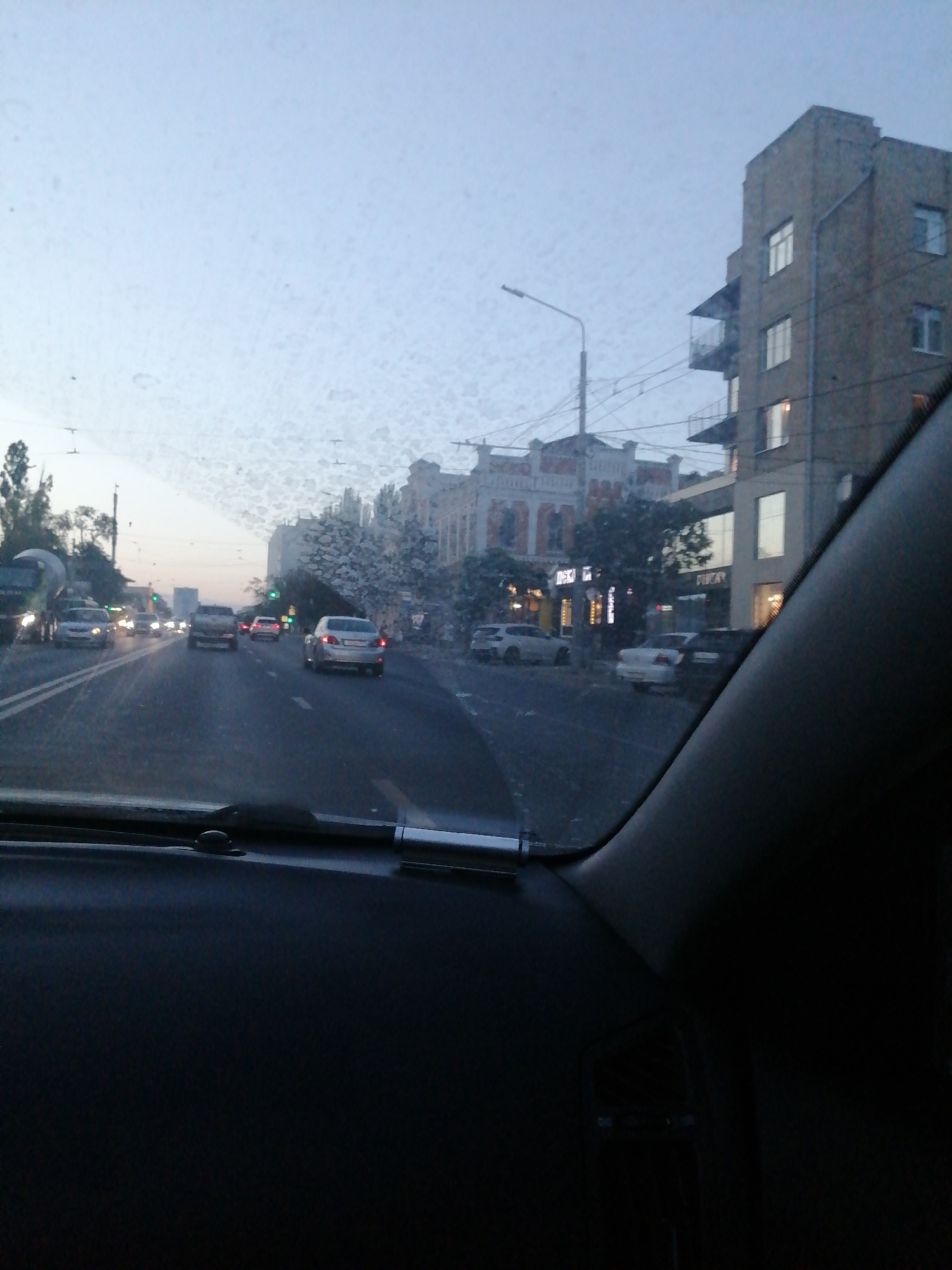 The image captures a view from inside a vehicle, looking out through a windshield with numerous water droplets and smudges. It depicts a street scene in Rostov-on-Don, Russia, likely during the daytime given the lighting conditions, though the sky appears overcast or hazy.
In the foreground, the dark interior of the car's dashboard is visible, partially obscuring the view. The windshield itself is the primary element in the mid-ground, marked by water droplets and streaks that distort the scene beyond.
The street is lined with buildings. On the right, a multi-story brick building with several windows and balconies is prominent. To the left and center, other buildings with architectural details are visible, some appearing older with ornate facades. Several cars are on the road, moving in the same direction as the vehicle from which the photo is taken. Traffic lights are also visible, some appearing to be red. Trees and streetlights line the sides of the road. The overall atmosphere suggests a somewhat muted urban environment, possibly a residential or mixed-use area.