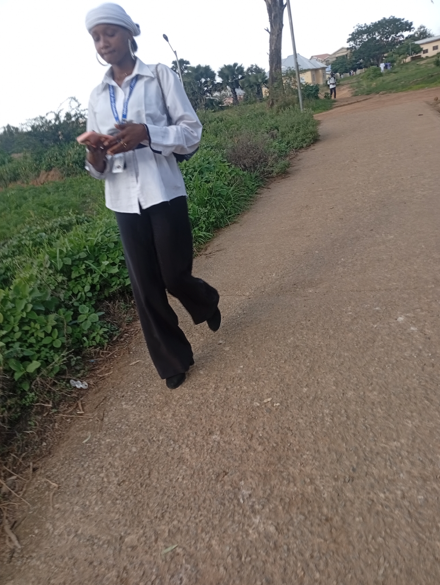 A full-shot photo shows a young woman with a headwrap and a white button-down shirt, black pants, and black shoes walking along a dirt path in what appears to be a rural or semi-rural area. The woman is looking down at her phone, which she holds in her right hand. She is wearing large hoop earrings and several rings. A blue lanyard with an ID card hangs around her neck. To the left of the path is dense green foliage. In the background, there are buildings, trees, and streetlights. The lighting suggests it is either early morning or late afternoon. The image is from Rafin Par, Nigeria.
