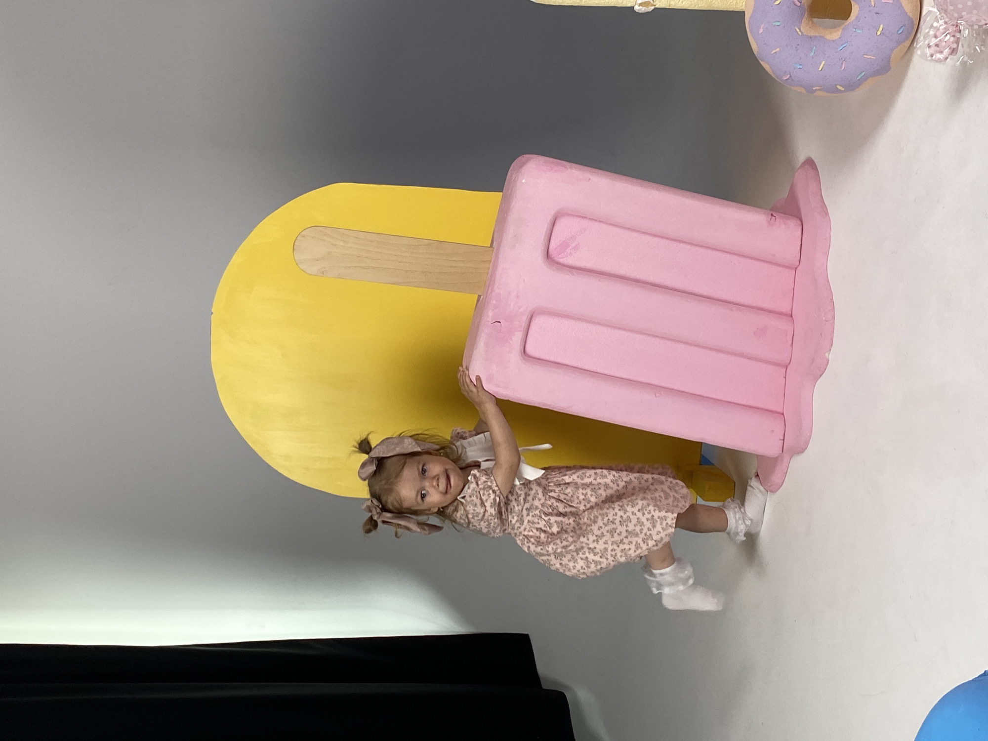 A young girl with blonde pigtails and a floral dress is posing with a large, pink, popsicle-shaped prop. She is holding onto the popsicle and looking at the camera with a happy expression. She is wearing white socks with ruffled trim. Behind her is a large yellow, popsicle-shaped prop and a purple donut prop. The background is a light gray wall. The scene appears to be indoors, possibly a photo studio, given the plain backdrop and controlled lighting. The overall mood is playful and cheerful. No text is visible in the image.