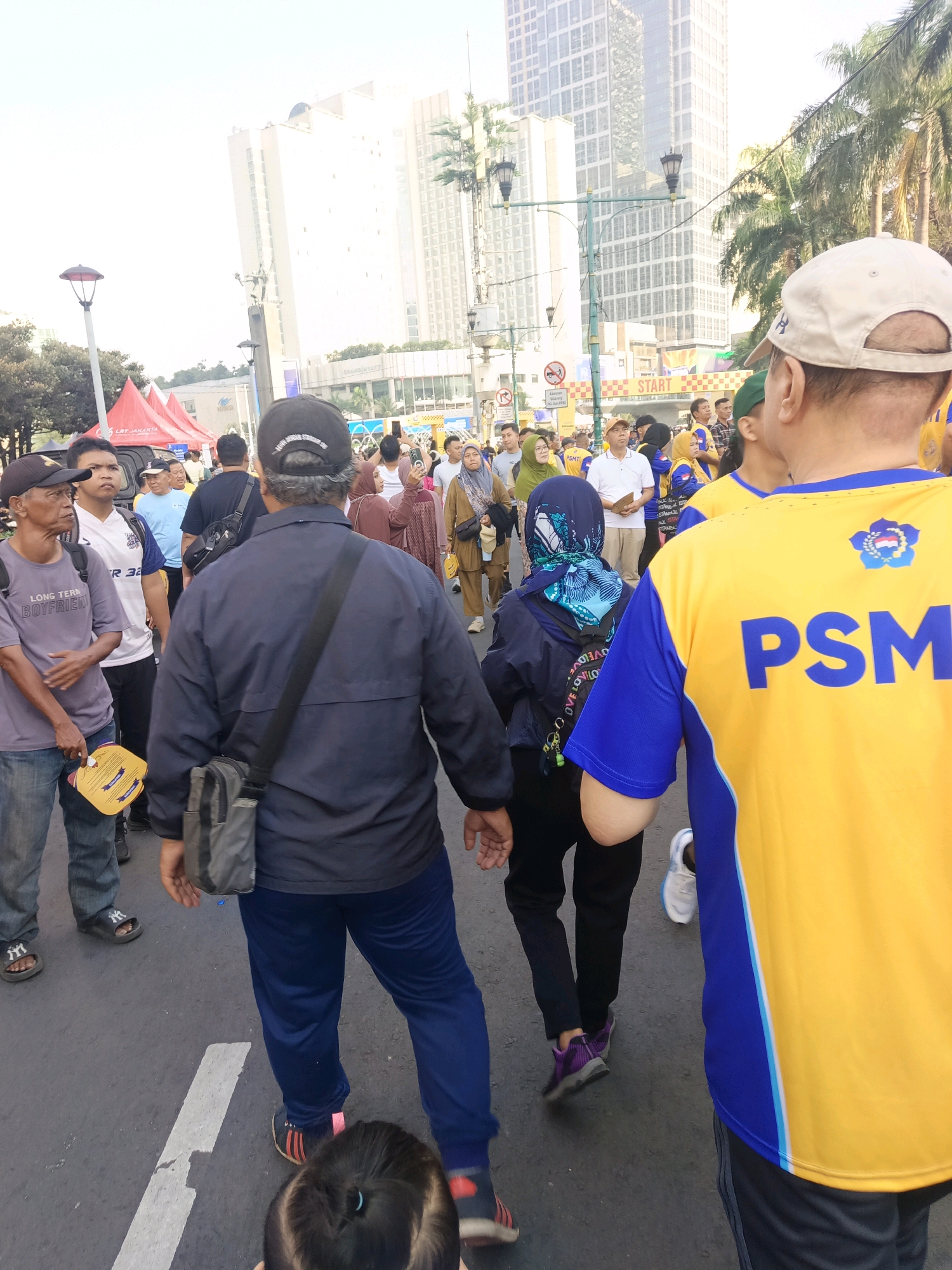 This is a photograph taken outdoors, in the city of Central Jakarta, Indonesia, during the daytime. The scene depicts a group of people walking on a paved road, likely participating in an event or activity. In the background, tall modern buildings dominate the skyline, with palm trees visible on the right side, suggesting a tropical urban environment.
In the foreground, several individuals are captured from the back or side. One person, on the far right, wears a bright yellow and blue shirt with the letters "PSM" prominently displayed. The person's back is towards the camera, and they are facing away from the viewer, walking into the distance. To their left, another individual is wearing a dark blue jacket over a dark shirt and dark pants. They are holding a gray bag slung over their shoulder and are walking in the same direction.
Further to the left, a man in a gray t-shirt and blue jeans is walking away from the camera. He is holding a yellow folded fan in his right hand. Next to him, a man in a white t-shirt with blue accents and a white cap is also walking. There are other people in the background, some wearing brightly colored headscarves and clothing, suggesting diversity within the group. Tents and signage are visible in the middle ground, indicating a possible organized event. The overall atmosphere appears to be lively and active.