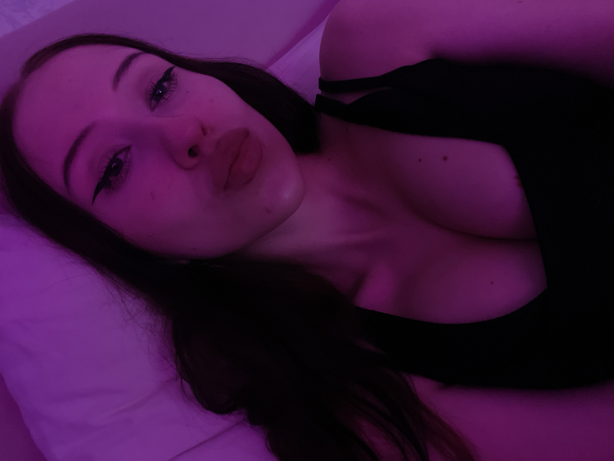 The image shows a close-up, high-angle selfie of a young woman with long, dark hair. She is wearing a black top and has eyeliner and lipstick on. Her lips are pursed, and she is looking towards the camera. The lighting is predominantly pinkish-purple, suggesting artificial lighting, possibly from a mood light or colored bulb. The background appears to be a bed with a white or light-colored surface, possibly a pillow or sheet. The overall setting is indoors, likely a bedroom. There is no indication of the city of Votkinsk, Russia, or any specific geographical location within it.