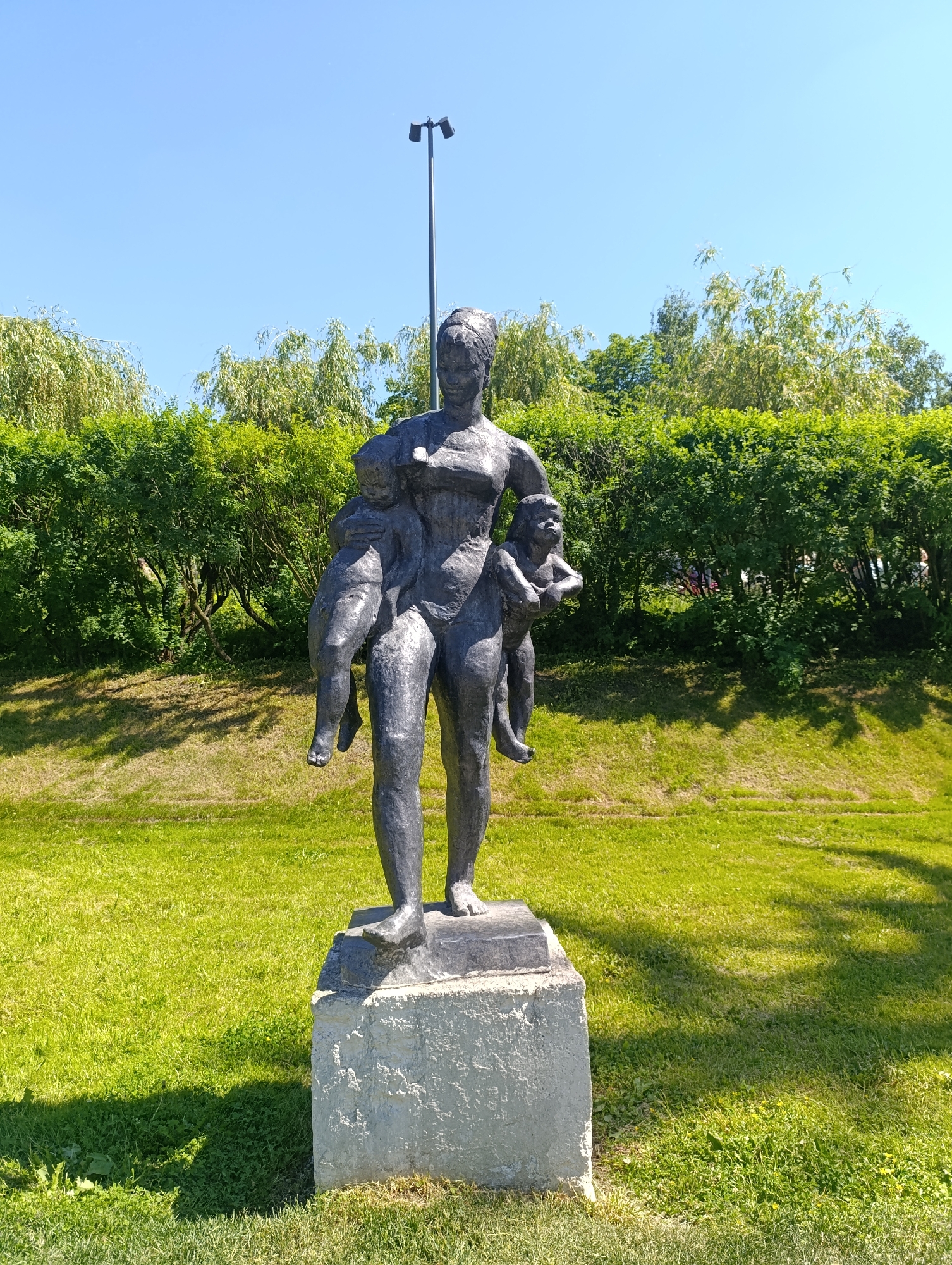 An outdoor, eye-level full shot captures a dark-colored statue depicting a standing woman holding two young children, one on each side. The statue rests on a light gray, rough-textured concrete pedestal positioned on a vibrant green grassy expanse. Bright sunlight illuminates the scene, casting distinct shadows from the statue onto the grass in the lower left. In the background, a dense line of various green bushes and trees, some with drooping foliage, extends horizontally across the frame. Above the vegetation, a clear, bright blue sky occupies the upper portion of the image. A tall, dark light pole with a bifurcated fixture is visible behind and to the right of the statue's head. The location is in the city of Unknown, Unknown.