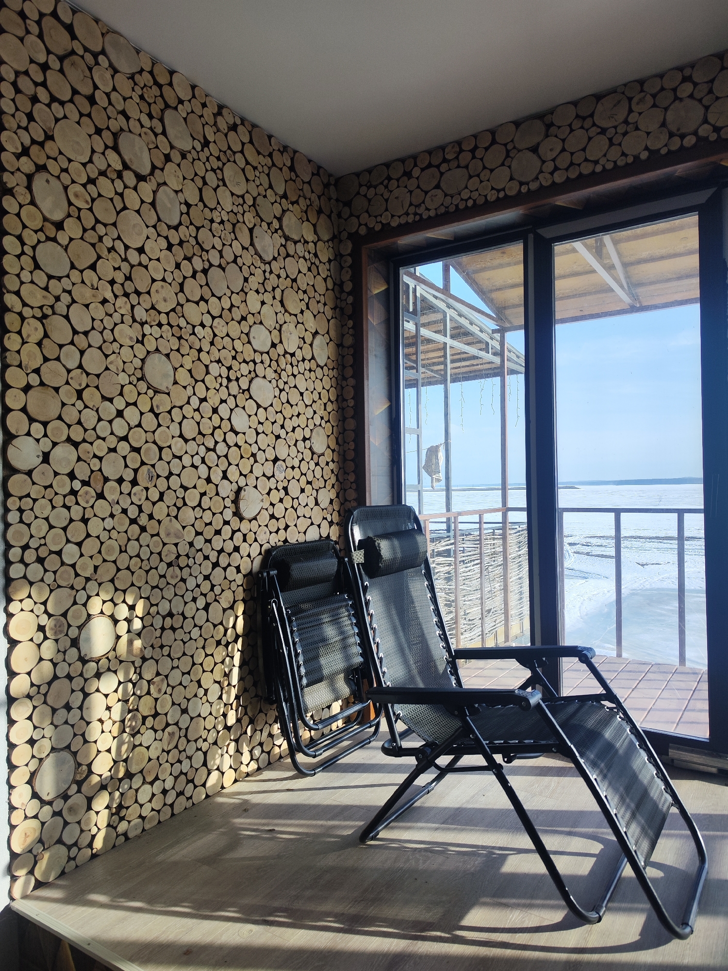 A room with a wall decoratively covered with cross-sections of small logs, two folding lounge chairs, and a large sliding glass door facing a snow-covered landscape. The scene is illuminated by sunlight streaming through the glass door. Outside, a building with visible metal framework and a snow-covered ground. The location may be Chaikovsky, Russia, based on context.