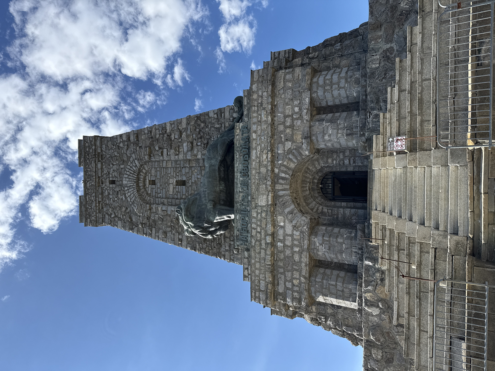The image shows the lower portion of the Shipka Memorial, a monument located in Shipka, Bulgaria. The monument is constructed of rough-hewn stone blocks and features a prominent bronze lion statue positioned above an arched entrance. A wide set of stone stairs leads up to the entrance, flanked by metal railings. The sky above is bright blue with scattered white clouds. Text is visible on the monument below the lion statue, appearing to be in Cyrillic script. A small sign on the stairs indicates "BILETI TICKETS" with an arrow and prohibition symbols.