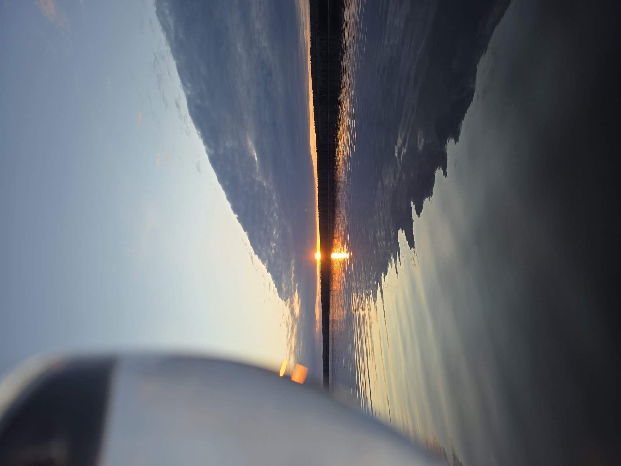 The image shows a sunset scene over water in Chaikovsky, Russia. The sun's reflection creates a bright line on the water's surface. A dark line of trees is visible on the horizon. The sky has a gradient of light blue to dark gray with clouds. The foreground includes a blurry white object.