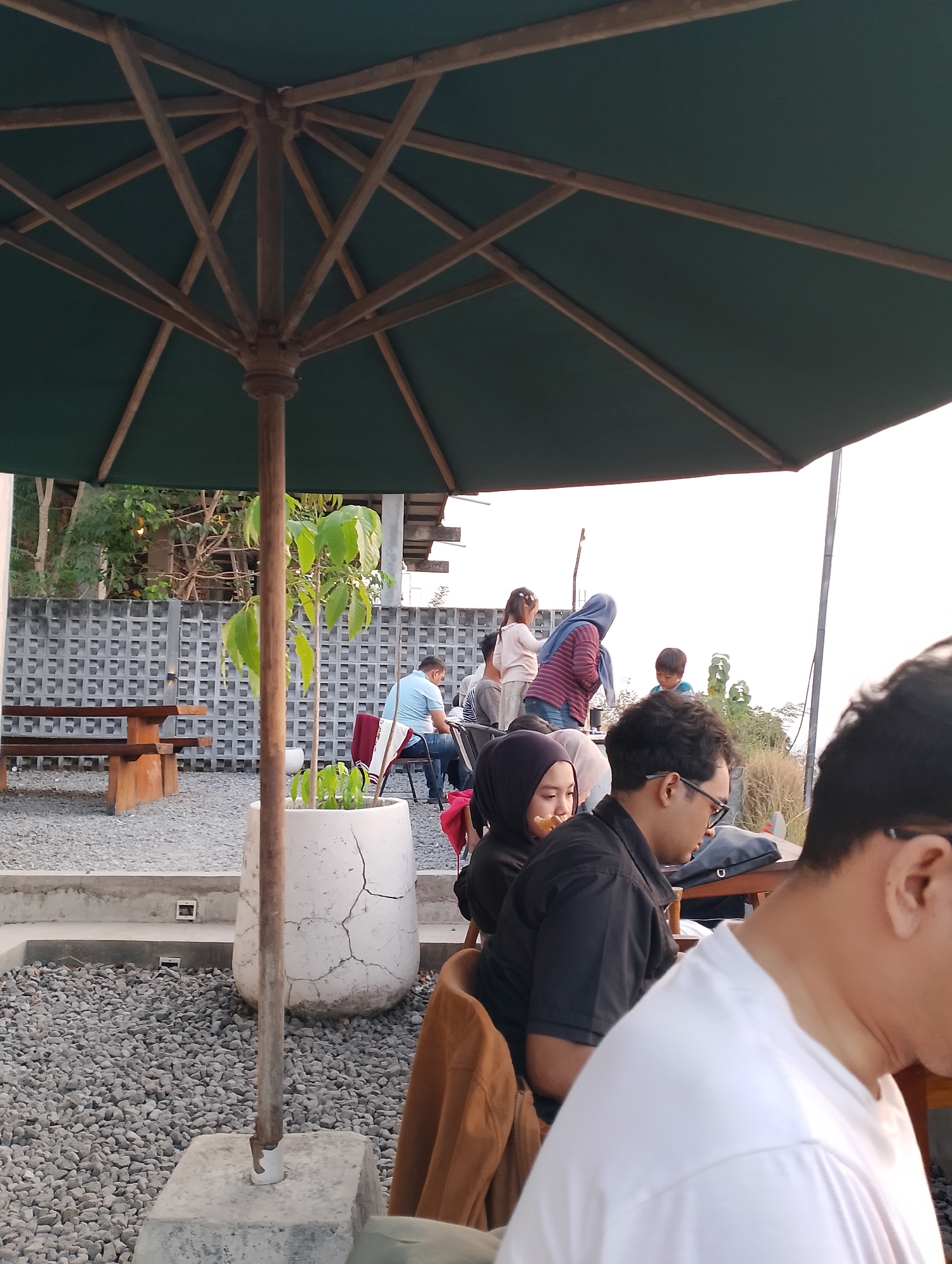 This image captures an outdoor seating area, likely at a cafe or restaurant, in Srimulyo, Indonesia. A large, dark green umbrella with a wooden frame provides shade over several patrons. In the foreground, a person wearing a white t-shirt is partially visible from the back. To their left, a man in a black polo shirt and glasses is seated, looking down. Beside him, a woman in a black hijab is also seated, with a piece of food near her mouth. Further back, a man in a blue shirt is seated at a table. In the background, other individuals are present, including a woman in a striped shirt and hijab, and two children. The ground is covered in gravel, and there are concrete elements and a wooden bench and table visible. A large, cracked white pot with a small plant stands near the center of the frame. The environment appears to be outdoors, with a low, patterned concrete wall and foliage visible in the distance.