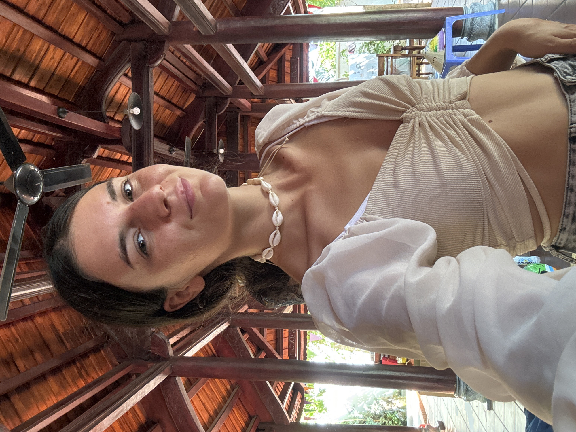 A young woman is taking a selfie in an outdoor, open-air structure with a wooden roof supported by pillars. She is wearing a beige, ruched crop top with sheer, voluminous sleeves and a white shell choker necklace. The woman has dark hair pulled back and is looking at the camera with a slight smile. The background shows the intricate wooden architecture of the building, along with some greenery and furniture. The setting appears to be a resort or a similar establishment in Mũi Né, Vietnam, likely a tourist area given the style of architecture and the conical hat on a blue stool in the background.