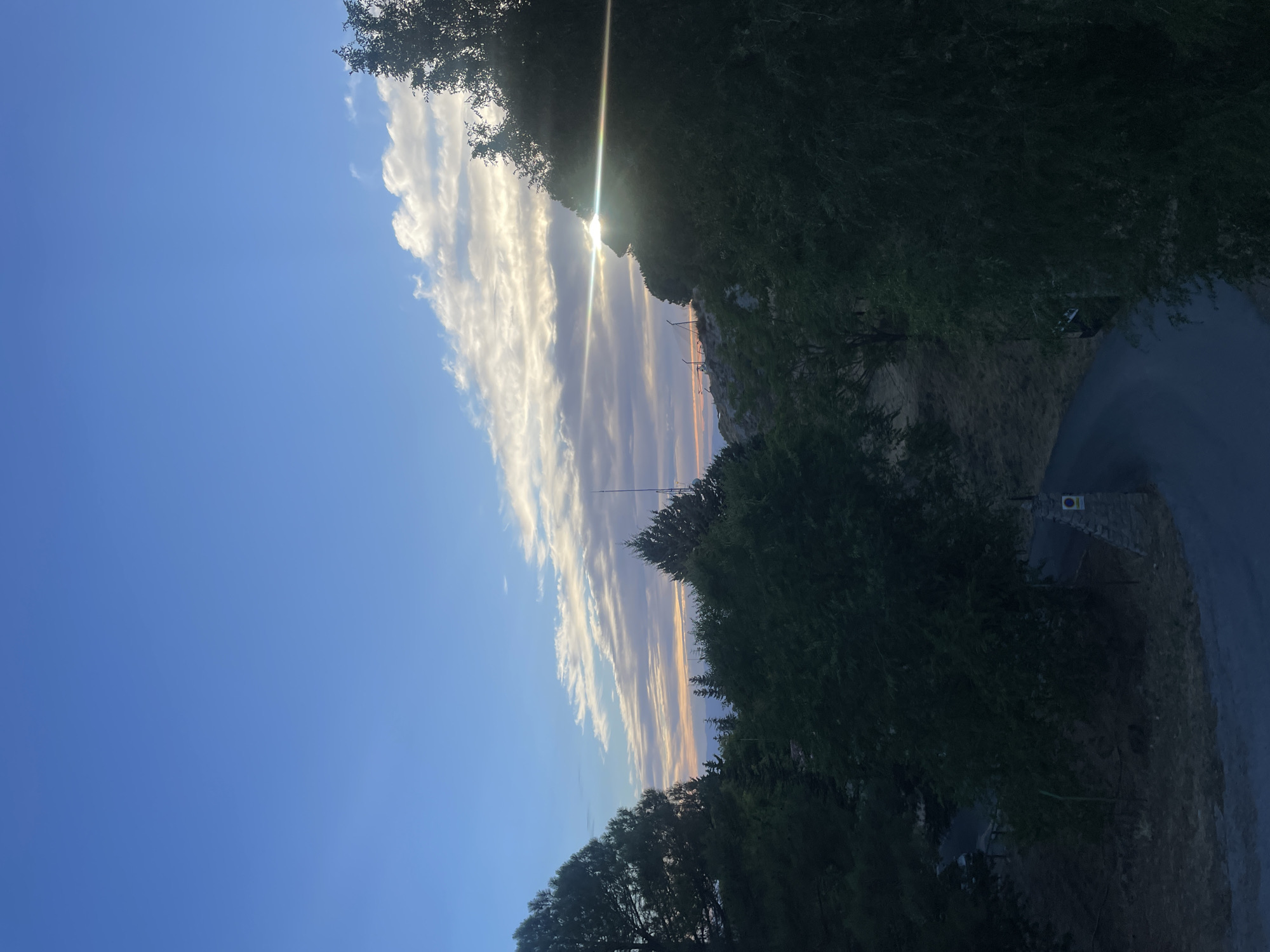 The image depicts a scenic view of the sky and landscape near Víznar, Spain, likely during sunset or sunrise. The sky is a vibrant blue, transitioning to softer hues near the horizon where wispy clouds are illuminated by the sun. The sun itself is partially obscured by trees and a ridge of land, casting a bright glare and lens flare across the scene. Dense foliage from various trees dominates the foreground and midground, with a winding road visible in the lower portion of the image. The road appears to be paved and leads through the terrain, flanked by dry, grassy slopes. A stone structure with a sign is situated next to the road. In the distance, a chain of hills or mountains is silhouetted against the colorful sky. The overall impression is one of natural beauty and tranquility, characteristic of a rural or mountainous area.