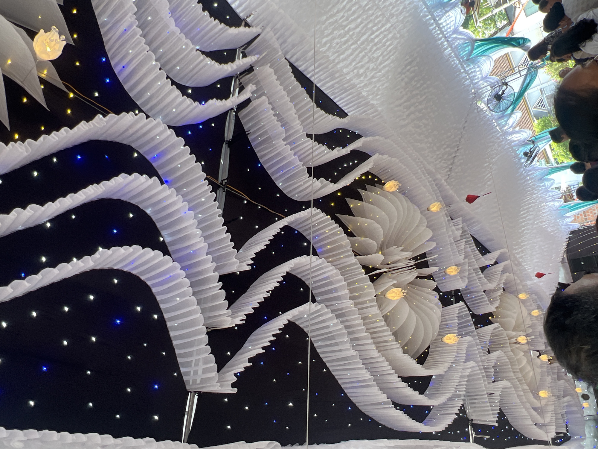 The image shows a street decoration in Bình An, Vietnam. The scene is an under-looking view of a street covered with overhead decorations. These consist of white ruffled streamers arranged in arches, spanning the street. The background above the streamers is black, dotted with twinkling blue and white lights, giving the impression of a night sky. Interspersed among the streamers are white flower-like structures, also illuminated. A few red flags are also visible among the white decorations. In the upper right of the image, people can be seen walking along the street, along with a bicycle. The light indicates it is daytime despite the decorations.