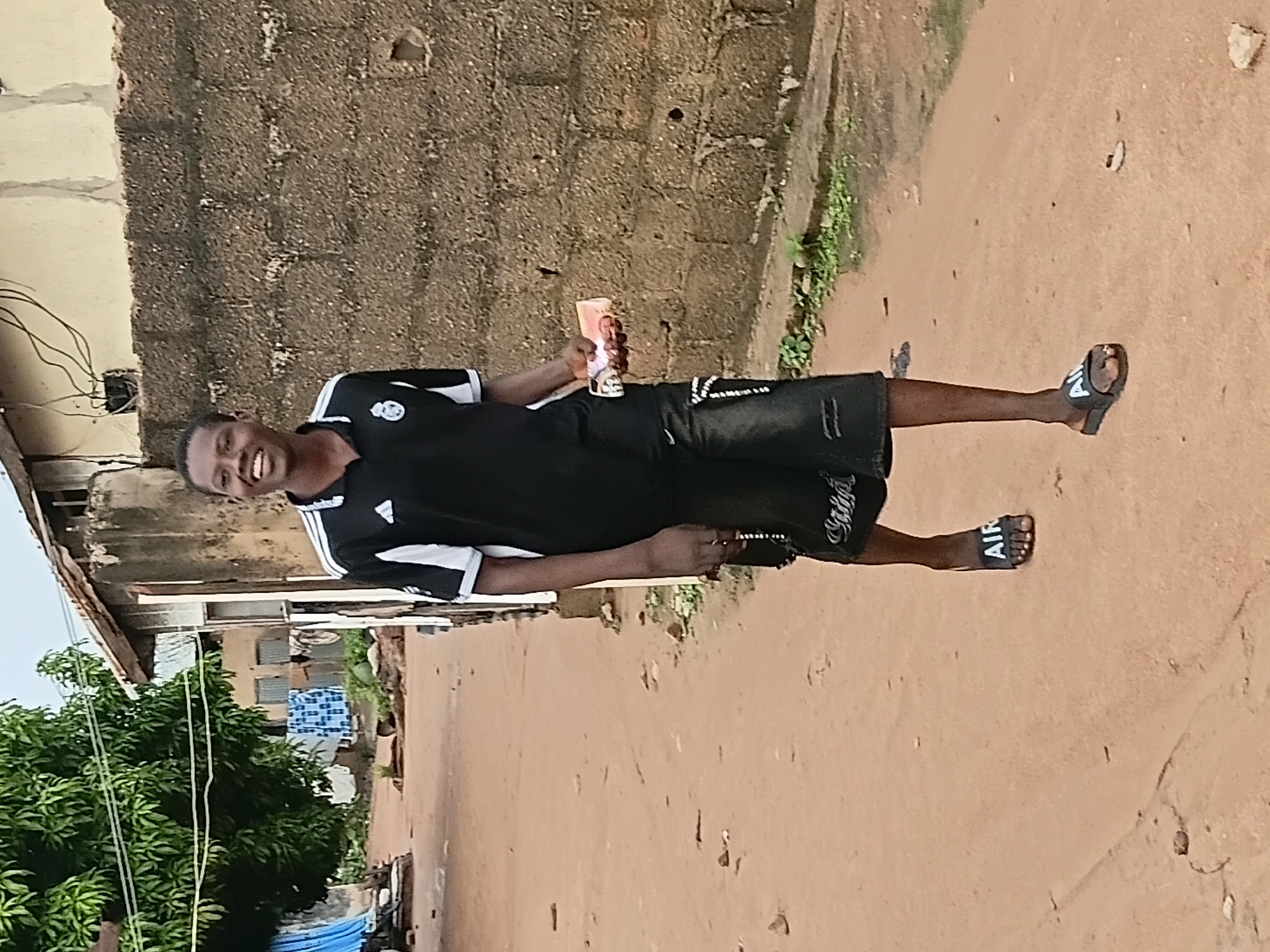 A young person with dark skin, wearing a black and white Adidas t-shirt and black shorts, walks along a dirt path in Jalingo, Nigeria. The person is smiling and holding a wad of cash in their right hand. The background shows a brick wall, some buildings with corrugated metal roofs, and a large tree. The path is dusty and uneven, with scattered rocks and debris. Laundry is hanging on a line in front of one of the buildings. The overall scene depicts a casual moment in an outdoor urban environment.