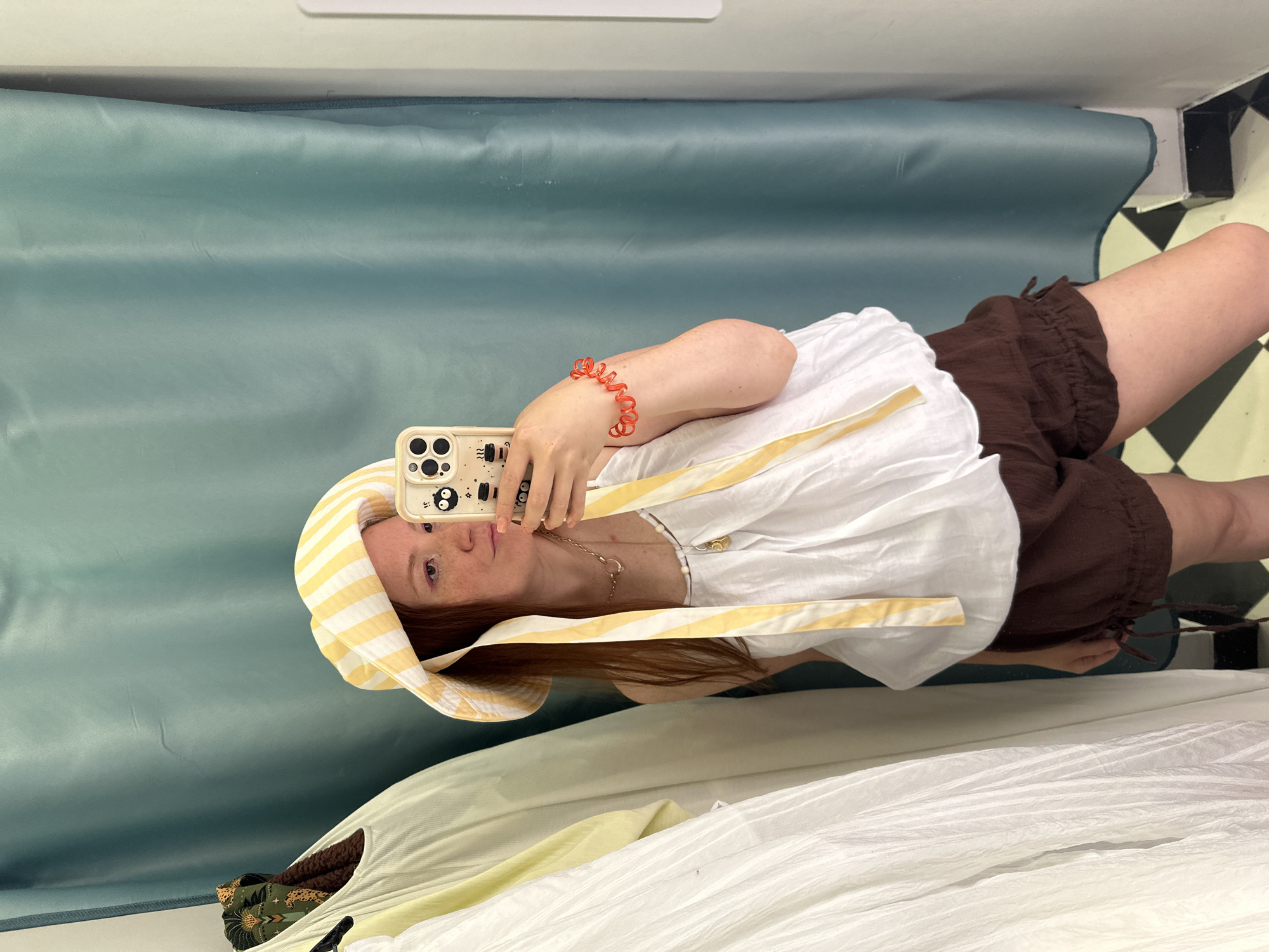 A woman is taking a selfie in what appears to be a fitting room. She is wearing a yellow and white striped hat, a white top with yellow straps, and brown shorts. She is holding a smartphone with a beige case featuring cartoon figures. On her wrist, she wears an orange bracelet.
The fitting room has a green curtain backdrop and a black and white checkered floor. Another garment, possibly a dress, is partially visible at the bottom of the frame. The location is unknown and there is no visual information that places the scene in Nizhny Novgorod, Russia.