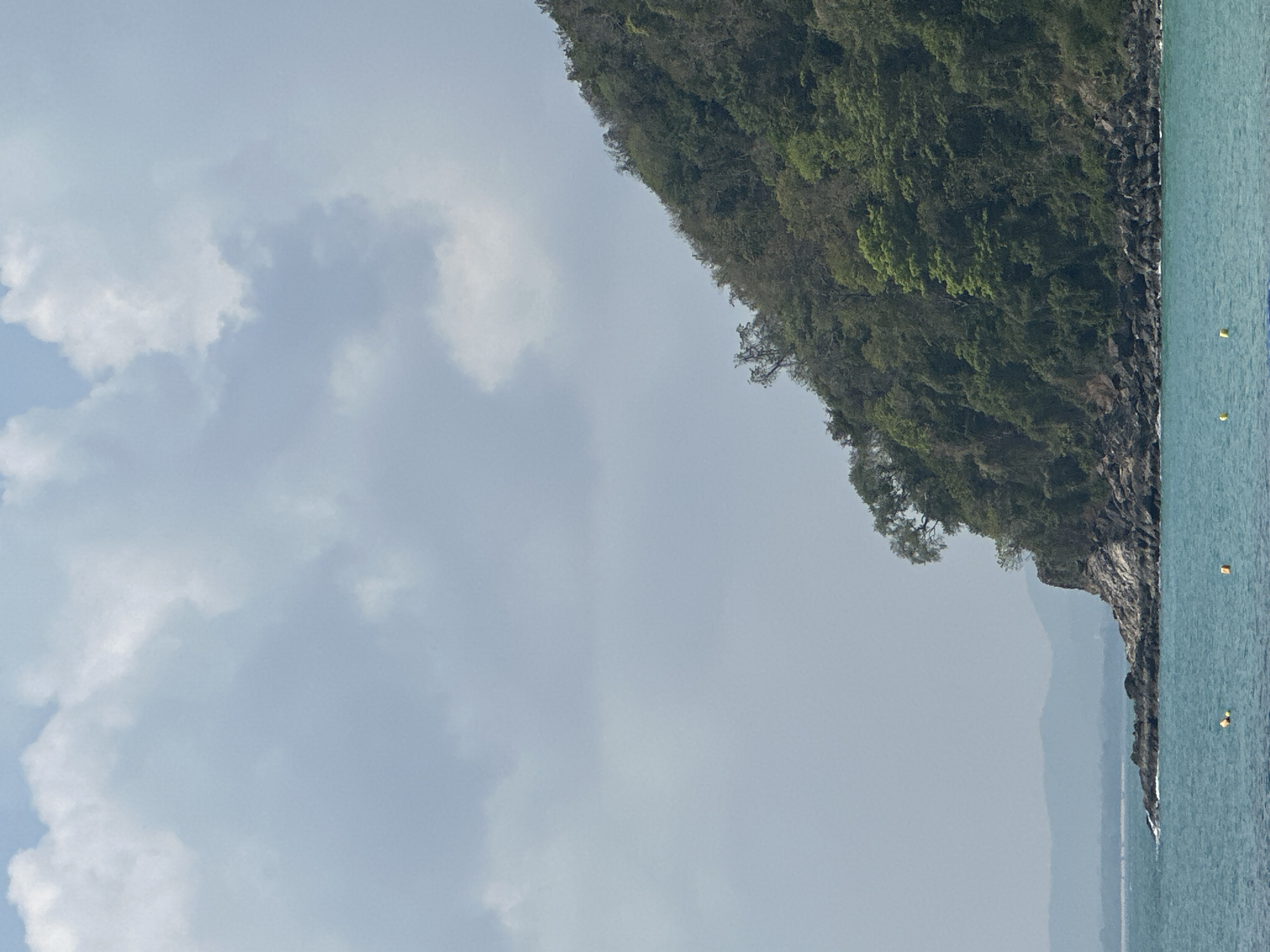 The image shows a coastal landscape in Ao Lukyon, Thailand. A lush, steep hillside covered in trees dominates the right side of the frame, bordering the sea. The sea appears calm and a few yellow buoys are visible. In the background, a lighter, indistinct mountain range is seen. The sky is overcast, with mostly grey clouds.
