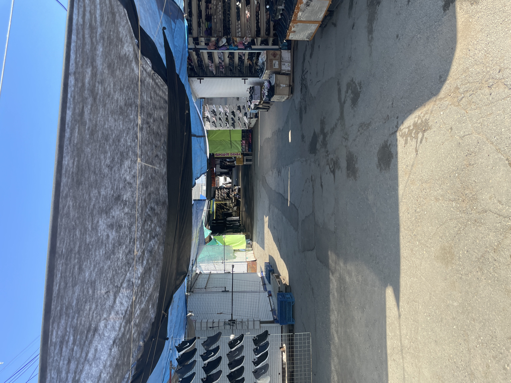 The image depicts a narrow, sunlit alley in Izhevsk, Russia, lined with market stalls. The alley's concrete floor is cracked in several places. The stalls are covered with black and blue tarps on the left and feature various merchandise displays on both sides, including footwear. Some stalls have white corrugated metal walls and display sneakers. Green and white lettering is visible on one stall. The sky is blue in the upper left corner. A shadow is cast on the right side of the alley. People are visible in the distance.