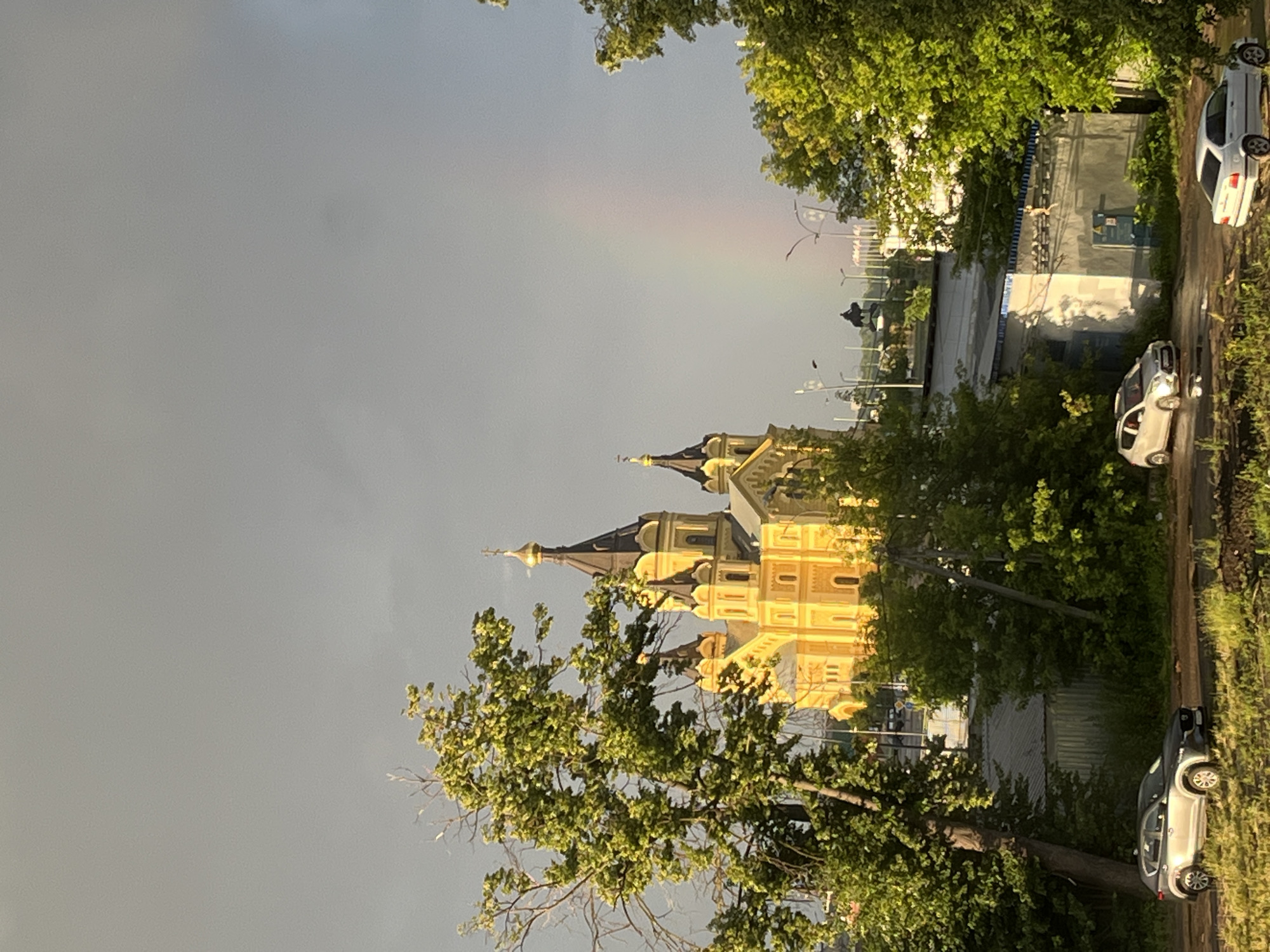 The image captures a city landscape in Nizhny Novgorod, Russia, featuring the Alexander Nevsky Cathedral prominently in the background. The cathedral, an iconic landmark, is bathed in the warm glow of sunlight, contrasting with the overcast, grey sky. A faint rainbow is visible in the sky, adding a touch of natural beauty to the urban scene. In the foreground, lush green trees frame the view, some with bare branches. Several cars are parked on a dirt road, with one silver SUV positioned on the left and two white cars further down the road. The ground appears wet, suggesting recent rain. The architecture of the cathedral, with its multiple domes and ornate details, is characteristic of Russian Orthodox churches. The presence of trees and the hint of a rainbow suggest a post-rain, possibly late afternoon or early evening, setting.