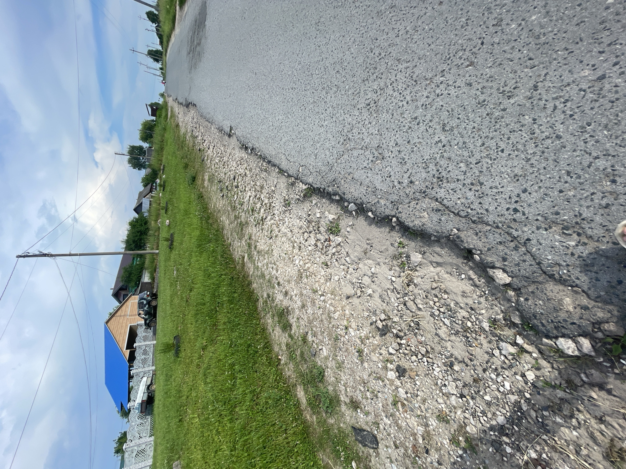 The image shows a rural road with damaged asphalt in the village of Krasnye Baki, Russia. A grassy embankment with loose gravel borders the right side of the road. In the background, several houses with blue and brown roofs are visible, along with a white fence, a trailer, and a green military-style vehicle. Power lines stretch across the sky, supported by wooden poles. The road itself appears to be a two-lane street, with one car visible in the distance. The overall scene depicts a typical village street in a summery setting with a cloudy sky.
