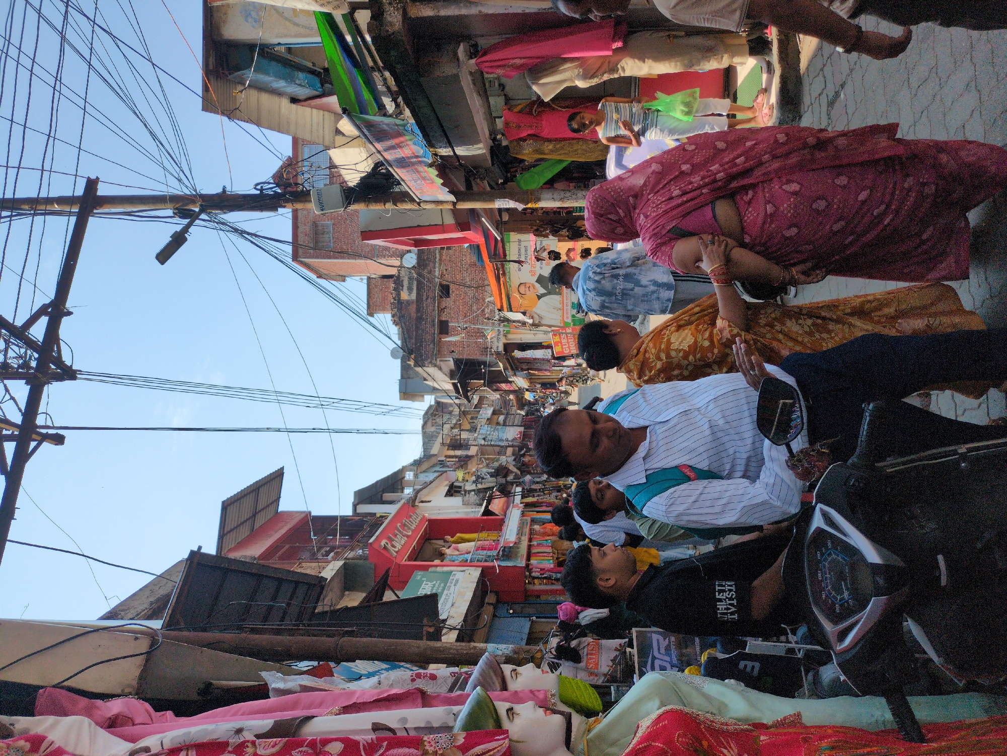 This is a medium shot, eye-level, outdoor photograph taken on a sunny day in Kangra, India. The scene depicts a bustling street market with a variety of stalls lining both sides of a narrow lane. In the foreground, a black scooter is parked on the cobblestone ground. To the left of the scooter, mannequins dressed in colorful clothing are displayed, suggesting a clothing store.
A man in a white and blue striped shirt, wearing a blue backpack, is standing in the middle of the frame. He appears to be interacting with another person who is partially visible to his right. To the left of the man in the striped shirt, a man in a black t-shirt with white lettering is standing. Further back, a woman in a bright pink sari is walking towards the right side of the image, with her arm around another person's arm.
The street is filled with people, indicating a lively market atmosphere. The buildings on either side of the street are two or three stories high, with a mix of architectural styles. Many of these buildings house shops, as evidenced by the displays of clothing and goods. Overhead, a dense network of electrical wires crisscrosses the sky, typical of many Indian urban environments. The sky is clear and blue. The overall impression is one of a vibrant and active marketplace.