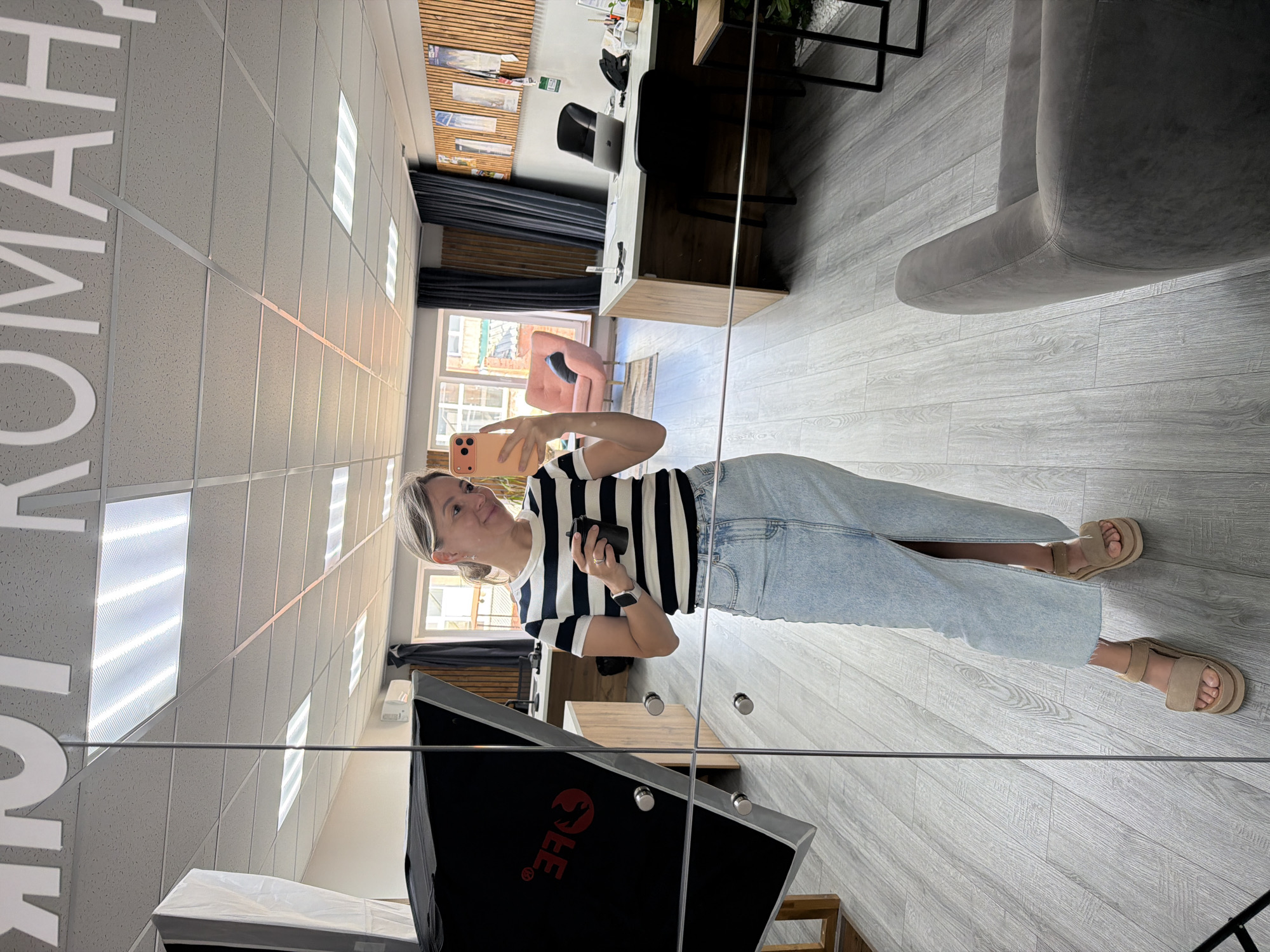 A woman is taking a mirror selfie in what appears to be an office or studio space. She is wearing a black and white striped t-shirt, light blue jeans, and tan sandals. She is holding a pink smartphone in her right hand and a black object, possibly a remote or a small device, in her left hand. She has blonde hair and is smiling.
The setting is a modern interior with a gray wooden floor and a white dropped ceiling with rectangular light fixtures. Large white letters are visible on the ceiling, partially obscured by a reflection. In the reflection, we can see a desk with a laptop, an office chair, and some decorative elements on the wall behind the desk, including wooden panels and framed pictures. A pink armchair is also visible in the reflection, as well as a window showing a brick building outside, suggesting an urban or suburban environment. A large black object with a red logo, possibly a piece of photography equipment, is present in the foreground of the reflection. The lighting suggests it is daytime. There is no visible text in the immediate scene, but the large white letters on the ceiling and the logo on the equipment are present in the reflection.