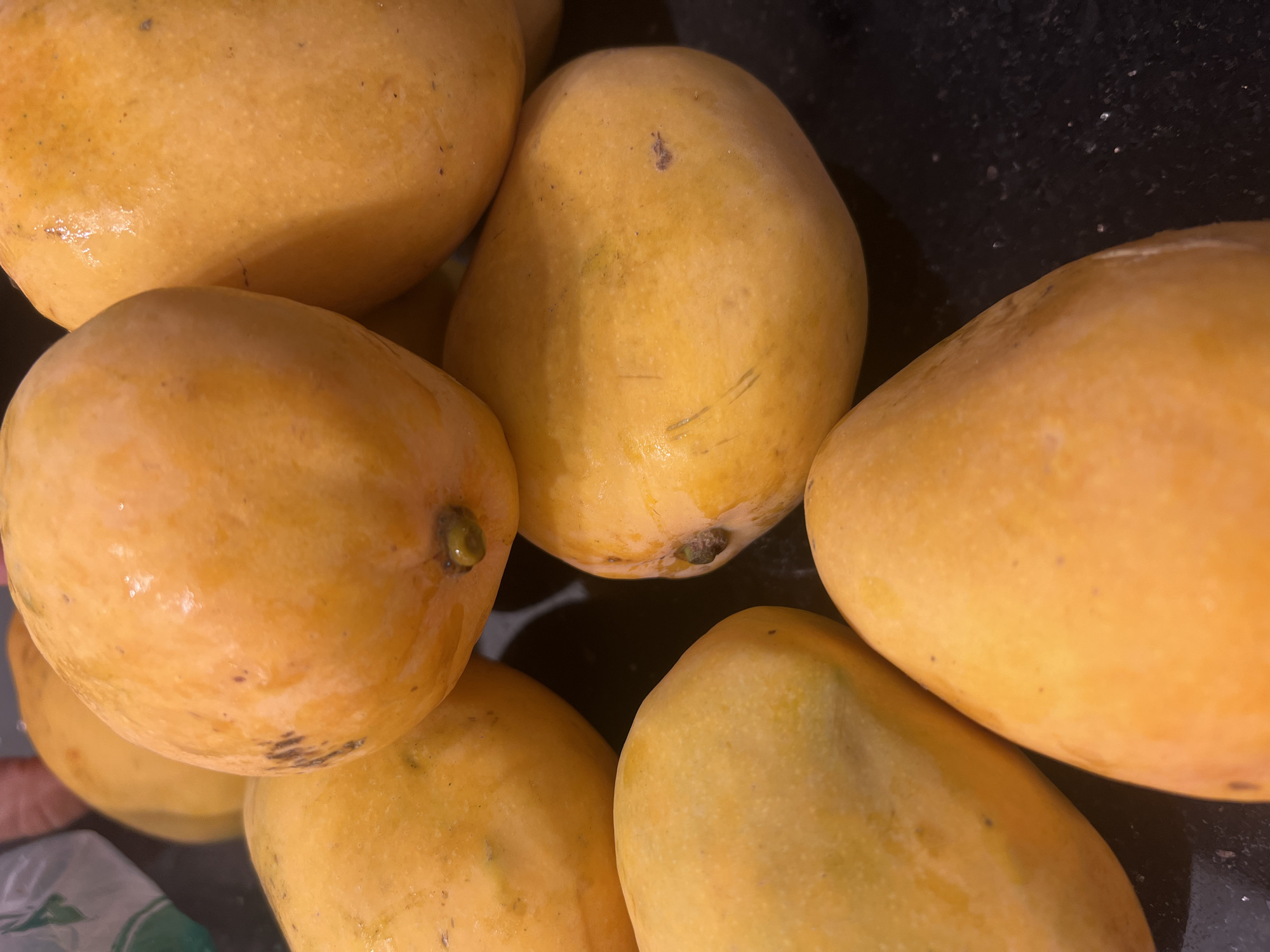 A close-up, top-down view shows a pile of ripe, yellow-orange mangoes arranged on a dark, speckled surface. Some of the mangoes have small brown spots or blemishes. The lighting is warm and slightly diffused, highlighting the glossy skin of the fruit and the subtle textures. In the upper left corner, a hint of a white plastic bag and a blurred pink object can be seen, suggesting the mangoes may have recently been purchased or are being stored. The scene is devoid of any human presence or activity. The dark surface appears to be a countertop or table. The image does not contain any elements that would place it specifically in Sharm El Sheikh, Egypt.