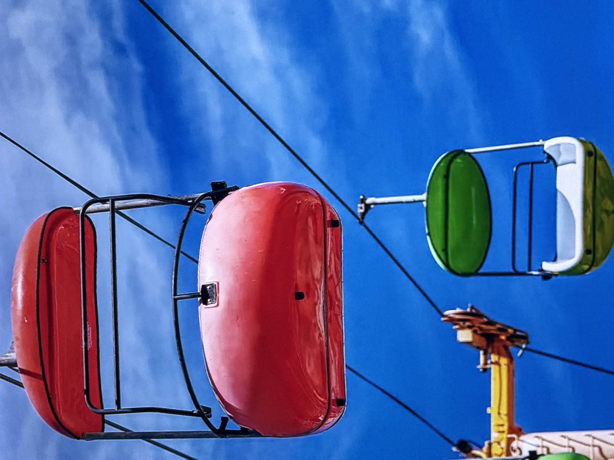 The image depicts two gondolas of a sky ride against a blue sky with wispy clouds. The gondola on the left is red and the gondola on the right is green and white. They are suspended by black cables. The scene is likely from a carnival or amusement park. The city and country are not discernible from the image.