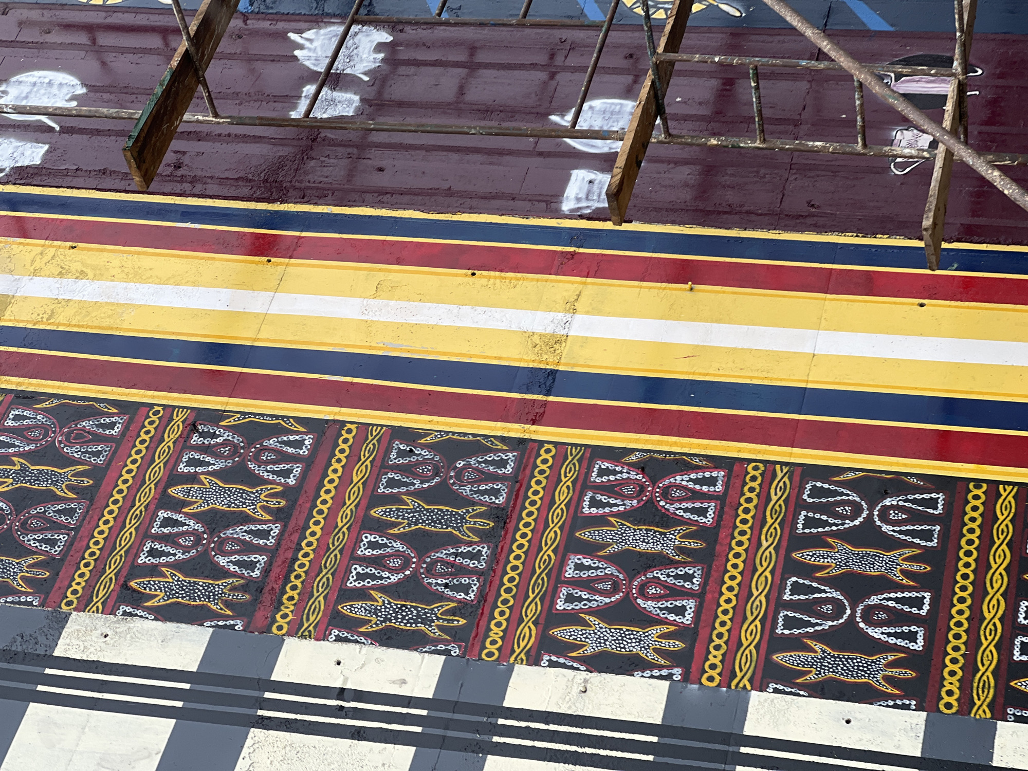 This is a close-up, eye-level, outdoor, medium shot of a mural painted on a weathered wall in Jalingo, Nigeria. The mural features a series of vertical stripes in red, yellow, white, and navy blue. To the left of the stripes, there is a section of the mural with a black background adorned with repeated patterns of stylized figures that resemble lizards and abstract shapes filled with white dots and red outlines. These patterns are bordered by bands of red and yellow wavy lines. To the right of the striped section, the wall is a deep maroon color with faint white outlines of what appear to be animal figures or abstract shapes. Scaffolding made of rusty metal pipes and wooden planks is positioned in front of the maroon section of the wall, partially obscuring the mural. The lighting suggests it is daytime, with some shadows cast from an unseen structure to the left of the frame.