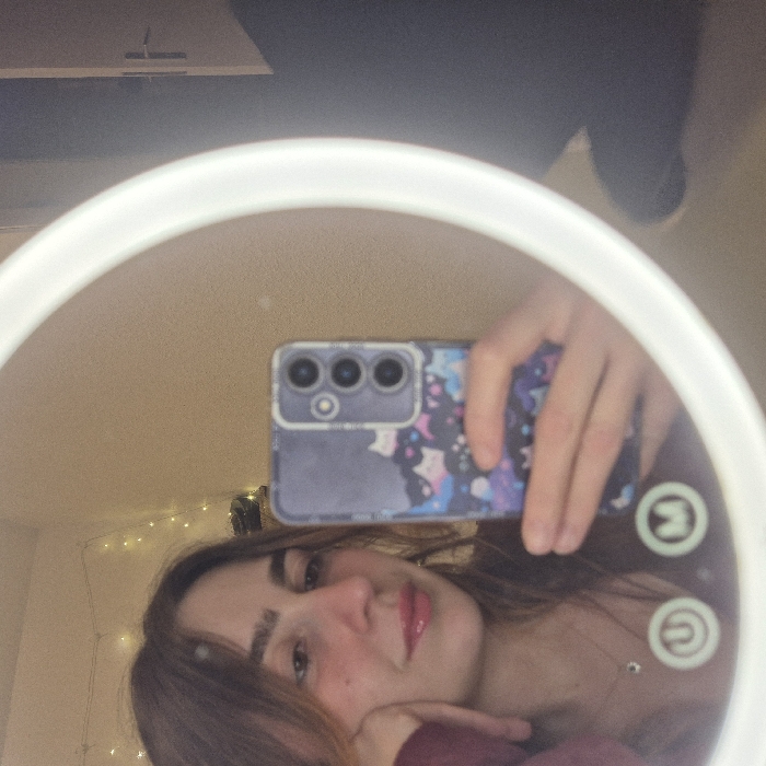 A selfie taken in a ring light mirror features a young woman with shoulder-length brown hair, fair skin, and glossy lips. Her phone, encased in a blue galaxy-themed case with three prominent camera lenses, is visible. The mirror reflects a glimpse of a room with faint string lights. The overall image has a warm, soft glow due to the ring light.