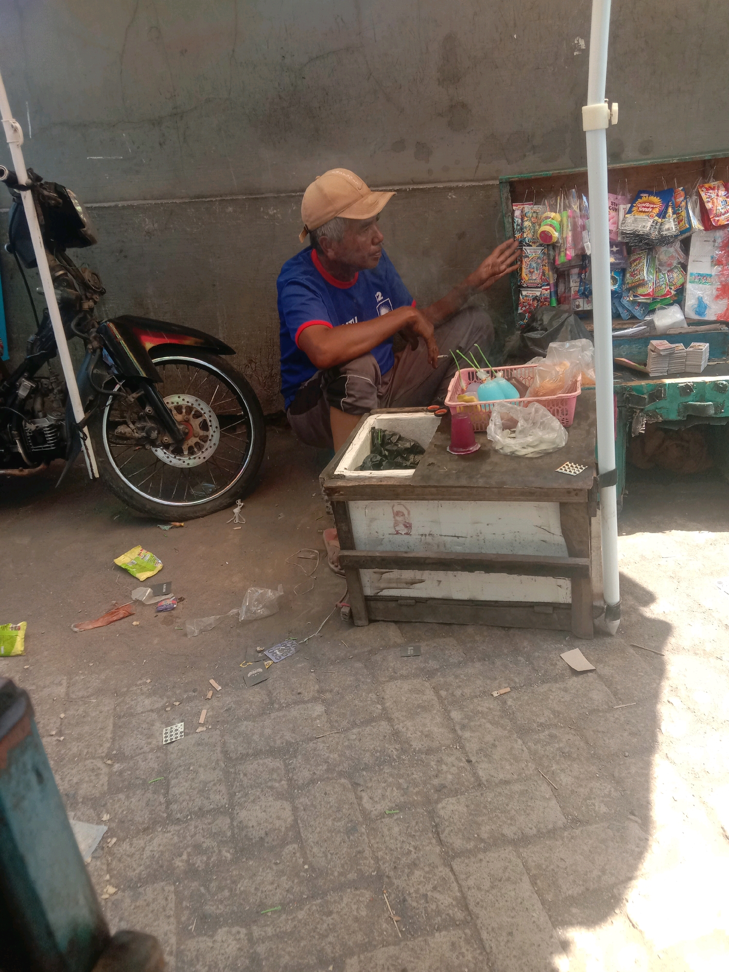 An elderly man with a light-colored cap sits on the ground next to a motorcycle and a vendor's stall in Karang Anyar, Indonesia. The man wears a blue shirt and dark pants. He appears to be gesturing with his right hand while looking to his left. The vendor's stall consists of a wooden table with various items for sale, including snacks in plastic packaging, drinks in a pink basket, and a cooler. The ground is paved with stone tiles and scattered with debris. A white pole, likely part of an umbrella or awning, stands to the right of the stall, casting a shadow. The background is a rough, gray wall.