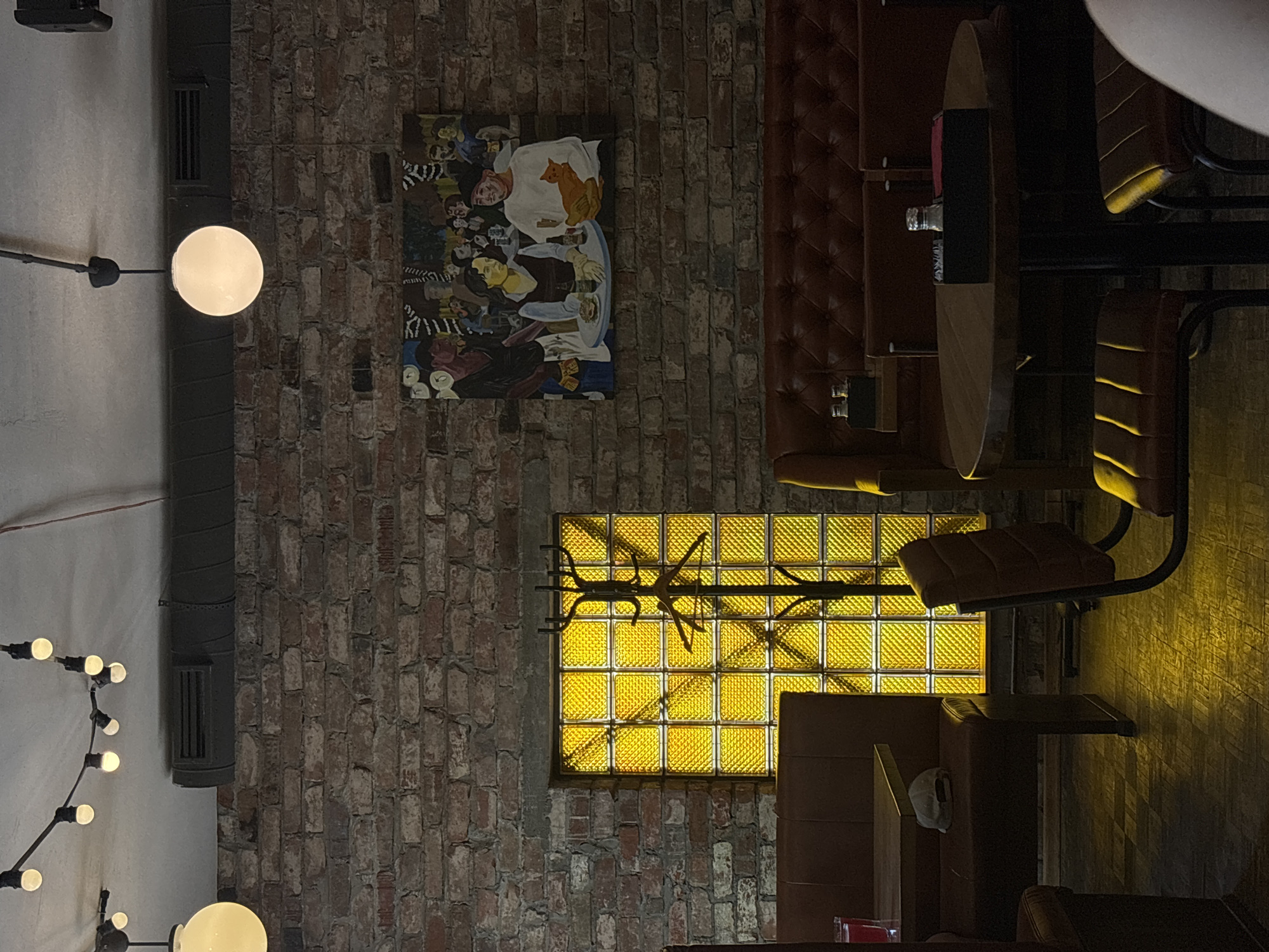 The image shows the interior of a restaurant or bar, with a brick wall, booth seating, and a round table. A painting hangs on the wall. A window with yellow glass bricks provides light. The location is likely Perm, Russia, based on contextual clues. No people are visible.