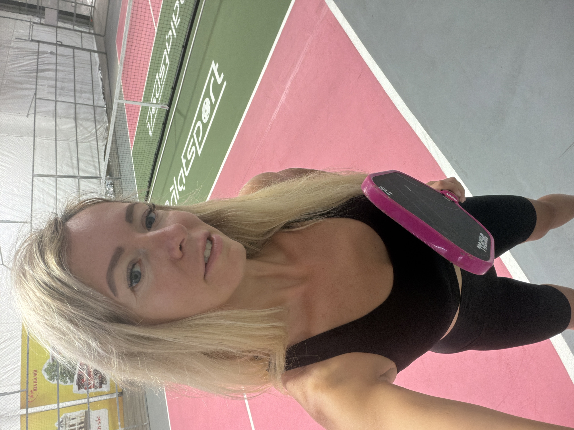 A blonde woman wearing a black sports bra and black cycling shorts is holding a pink pickleball paddle and looking at the camera. She is standing on a pickleball court, which has pink and green surfaces with white lines. In the background, there is a net, a fence, and a banner with Vietnamese text and images. The location is identified as a pickleball court in Da Lat, Vietnam.