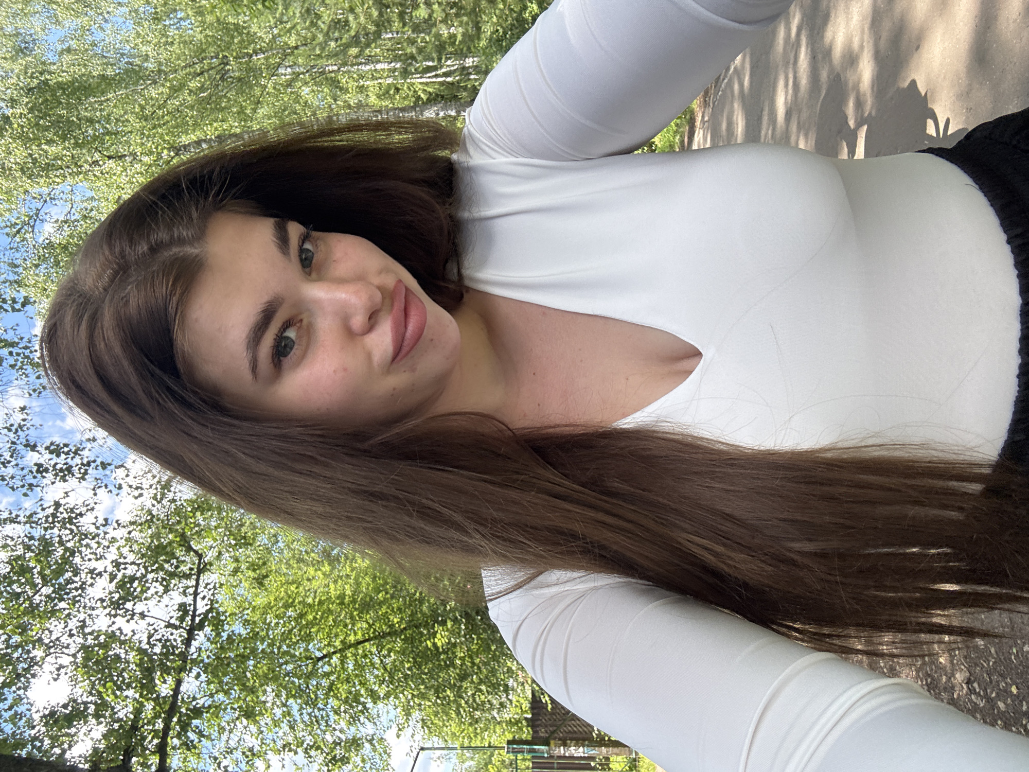 A young woman with long, dark brown hair stands outdoors, taking a selfie. She is wearing a white long-sleeved shirt with a V-neck and black pants. The background features lush green trees and a bright blue sky with white clouds. The setting appears to be a park or a natural area within the city of Meshchera, Russia, with a path visible in the foreground. Sunlight filters through the leaves of the trees, casting dappled shadows on the ground.
