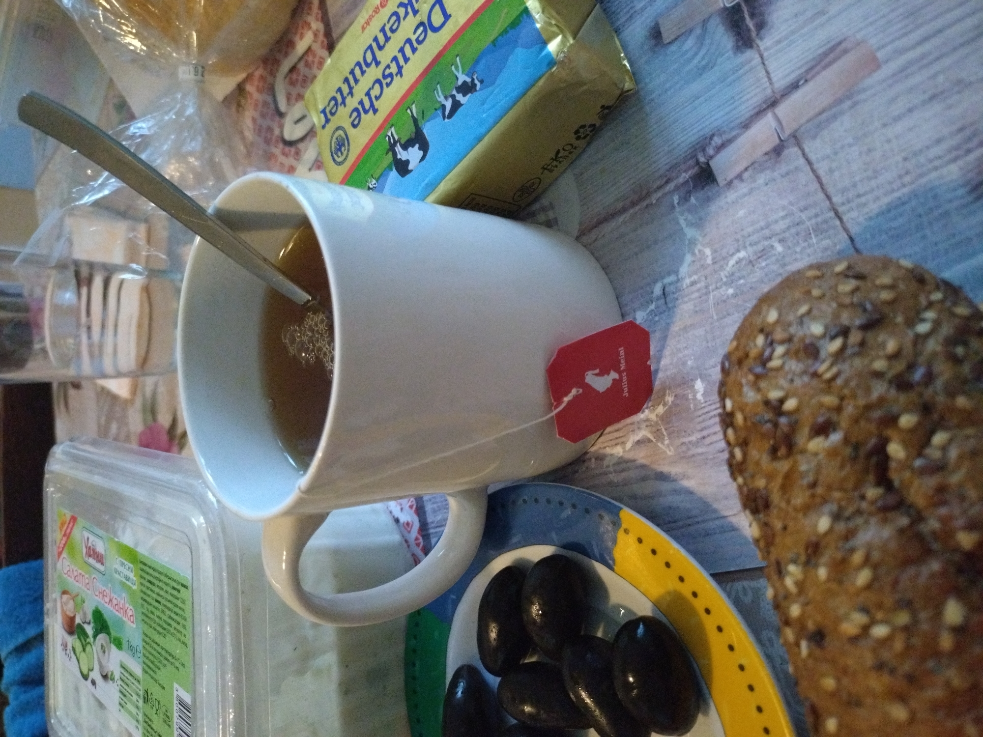 The image depicts a breakfast setting indoors. A large white mug filled with tea, a spoon inside, and a Julius Meinl tea bag tag are visible. Next to it, there is a blue and yellow plate containing black olives. A seeded roll of bread is also present. A package of "Deutsche Markenbutter" (German Butter) sits nearby. A plastic container labeled "Салата Снежанка" (Snezhanka Salad) is also in view. The scene is laid out on a surface with a wooden plank pattern. The precise location within Stara Zagora, Bulgaria cannot be determined solely from the image. No people or interactions are visible. The focus is primarily on food and beverage items prepared for consumption.