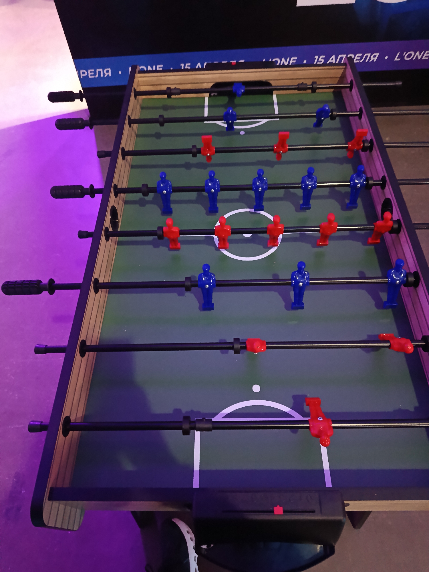 The image shows a foosball table with blue and red players. The table has a green playing surface with white lines marking the field. The table is located in an area with purple lighting, likely indoors, in the city of Unknown, Unknown.