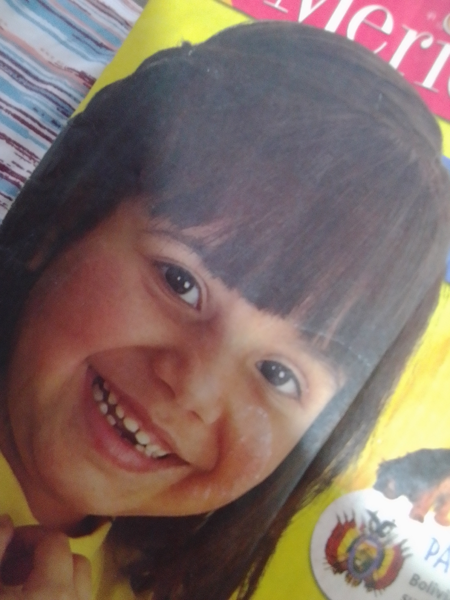 A close-up, slightly blurred image features a young child's face, tilted to the left, with dark hair and bangs. The child's eyes are open, and they have a wide, toothy grin. The background is out of focus, showing a colorful striped pattern and a portion of a yellow surface with text and an emblem. The emblem appears to be the coat of arms of Bolivia. The location in the city of La Tagua, Venezuela, is not discernible from the image.