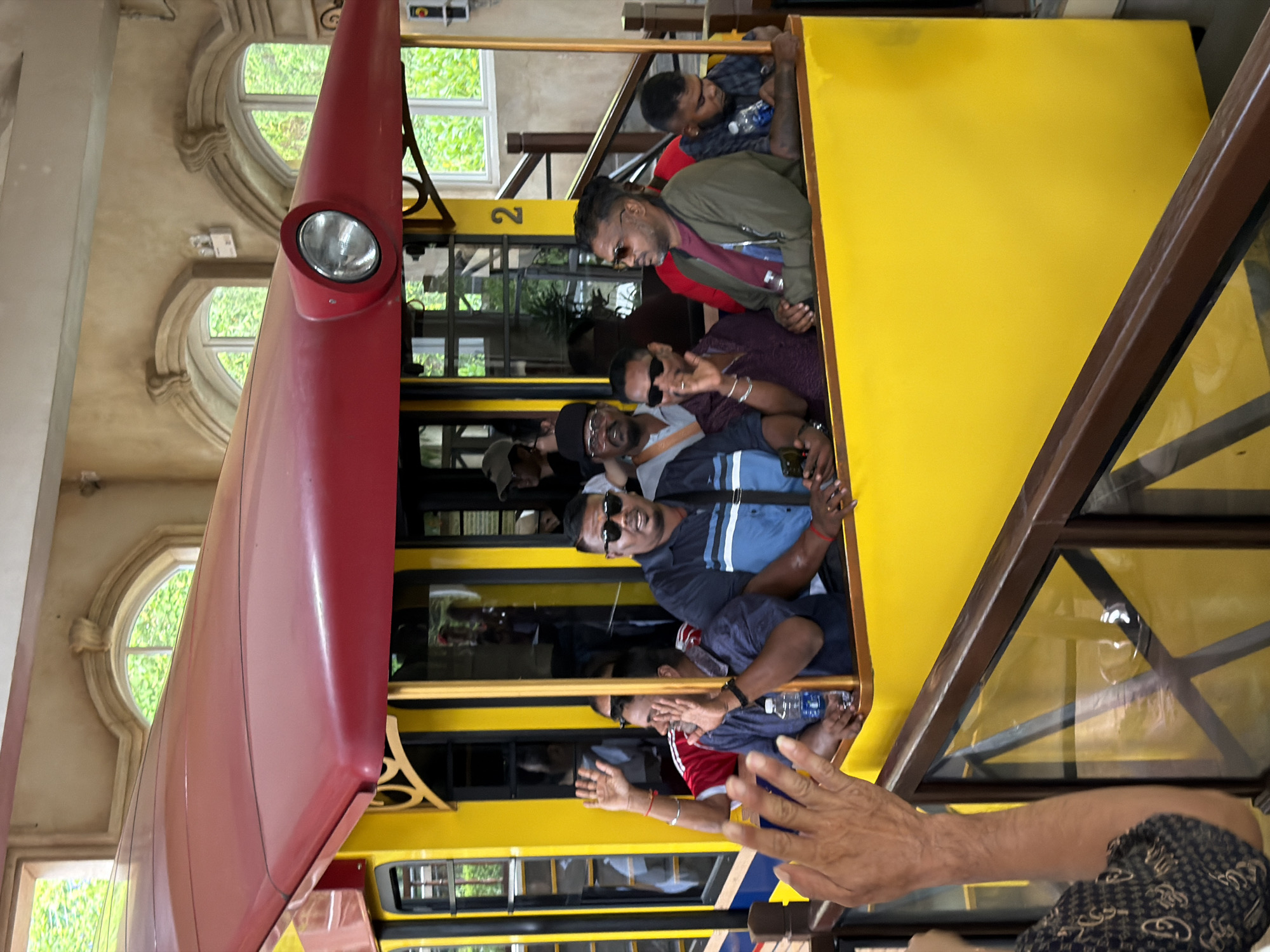 In the city of Da Nang, Vietnam, at Ba Na Hills, a group of people are riding in a bright yellow cable car. The cable car has a red roof and large windows. Inside, passengers are seated and looking out. One person in the foreground is waving with their left hand. Several passengers are wearing sunglasses. The cable car is moving along tracks. The environment outside the cable car appears to be lush and green, with trees visible through the windows. The architecture of the building in the background suggests a themed attraction. The number "2" is visible on the side of the cable car.