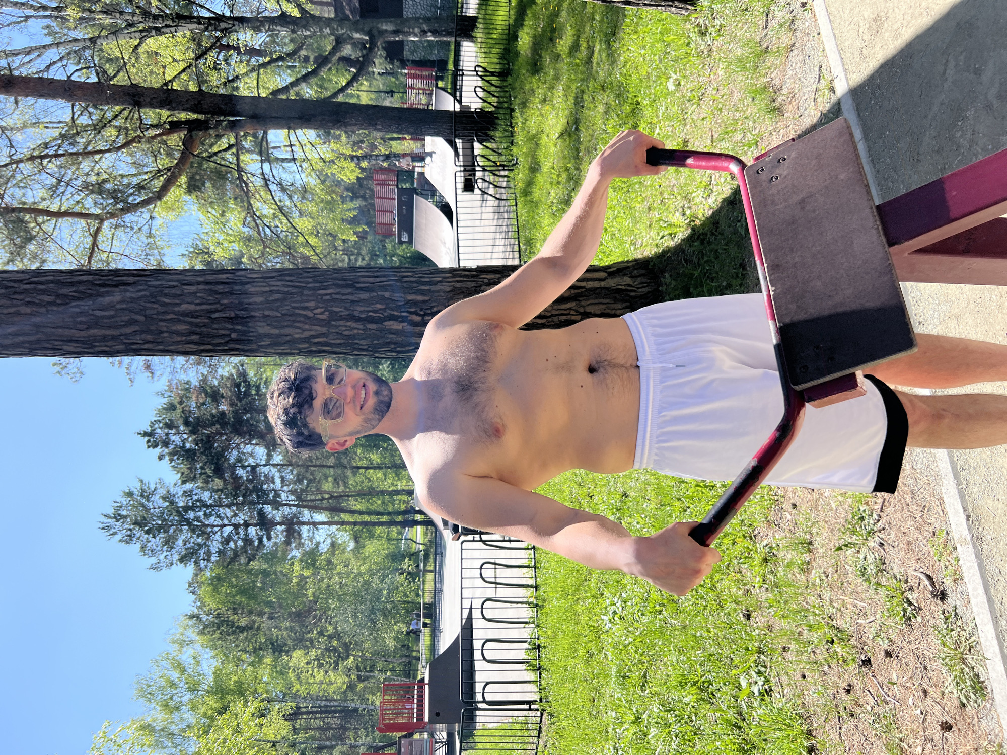 A man with dark hair and a bare chest is standing in an outdoor workout area, holding onto a piece of exercise equipment. He is wearing white shorts with a black stripe on the side. The area appears to be a park or recreational space with trees and green grass visible in the background. A bicycle rack and a skatepark can also be seen in the background. Based on the location, it is likely that this image was taken in Novosibirsk, Russia.