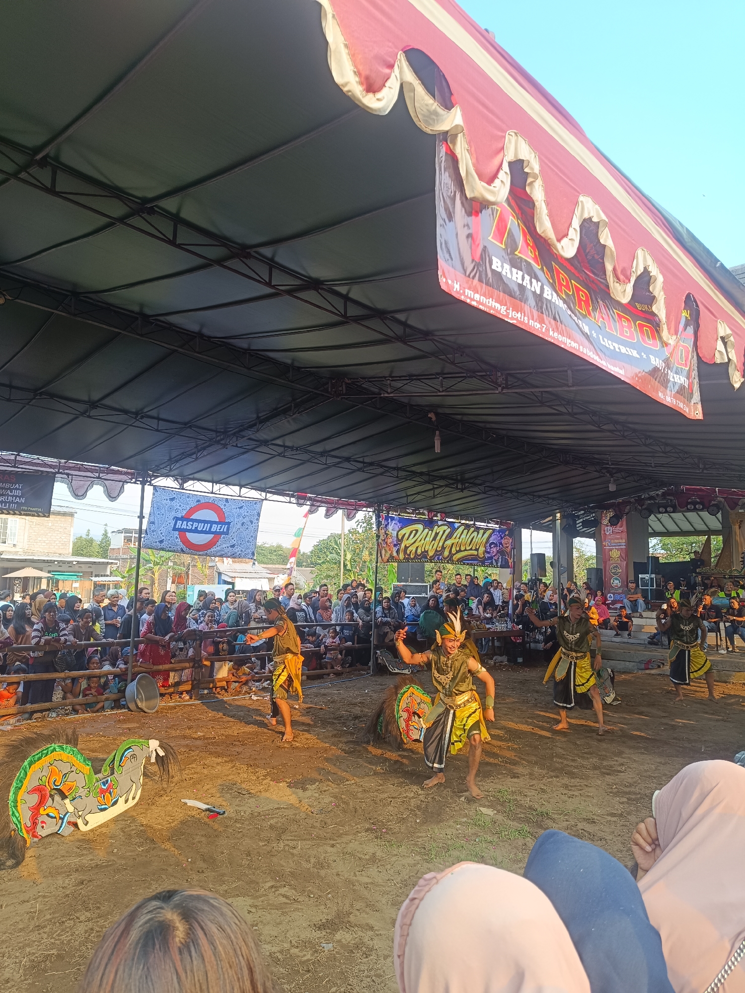 The image depicts an outdoor performance taking place under a large tent structure in Jetis, Indonesia. In the foreground, the backs of several audience members are visible, facing the performance area. The performance itself features several individuals in traditional costumes dancing on a dirt-covered stage. One performer in the center is wearing a green and yellow outfit with a headdress, while others are similarly attired. To the left of the performers, a colorful prop resembling a horse's head, decorated with intricate patterns, is placed on the ground. The background reveals a large crowd of spectators gathered behind a barrier, watching the event. Banners hang in the background, one with the text "RASPUJI BEN" and another with "PANJI ANOM". The overall scene suggests a cultural festival or traditional performance.