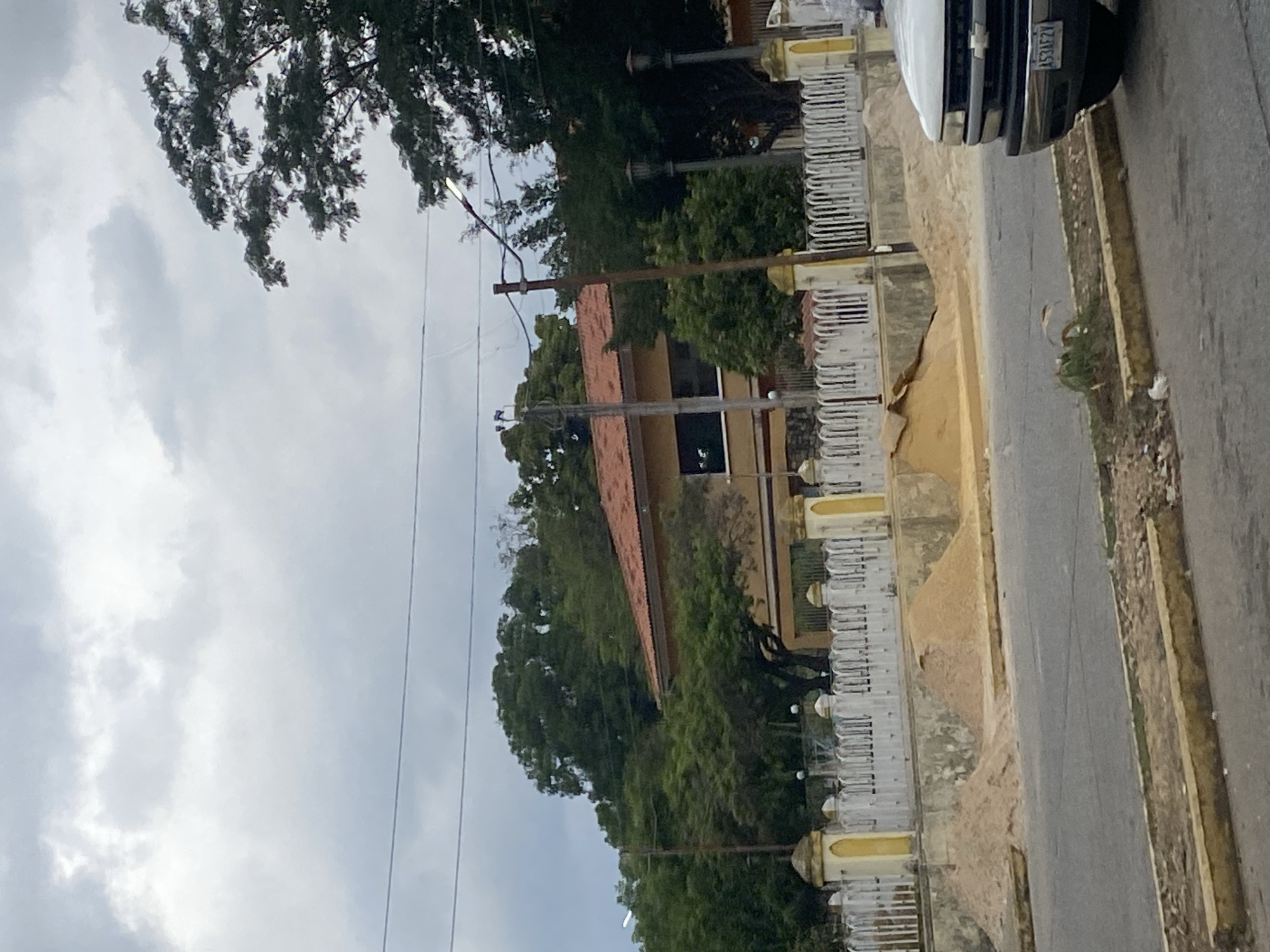 The image depicts a street scene in Tinaquillo, Venezuela, with a building set back from the road behind a white fence with yellow trim. The building has a red tile roof and tan walls. Trees and other foliage are visible around the building. A road runs alongside the fence with a section of curb visible. A white vehicle with a visible license plate is parked on the road, partly obscuring the fence. Overhead, there are power lines and tree branches against a cloudy sky.