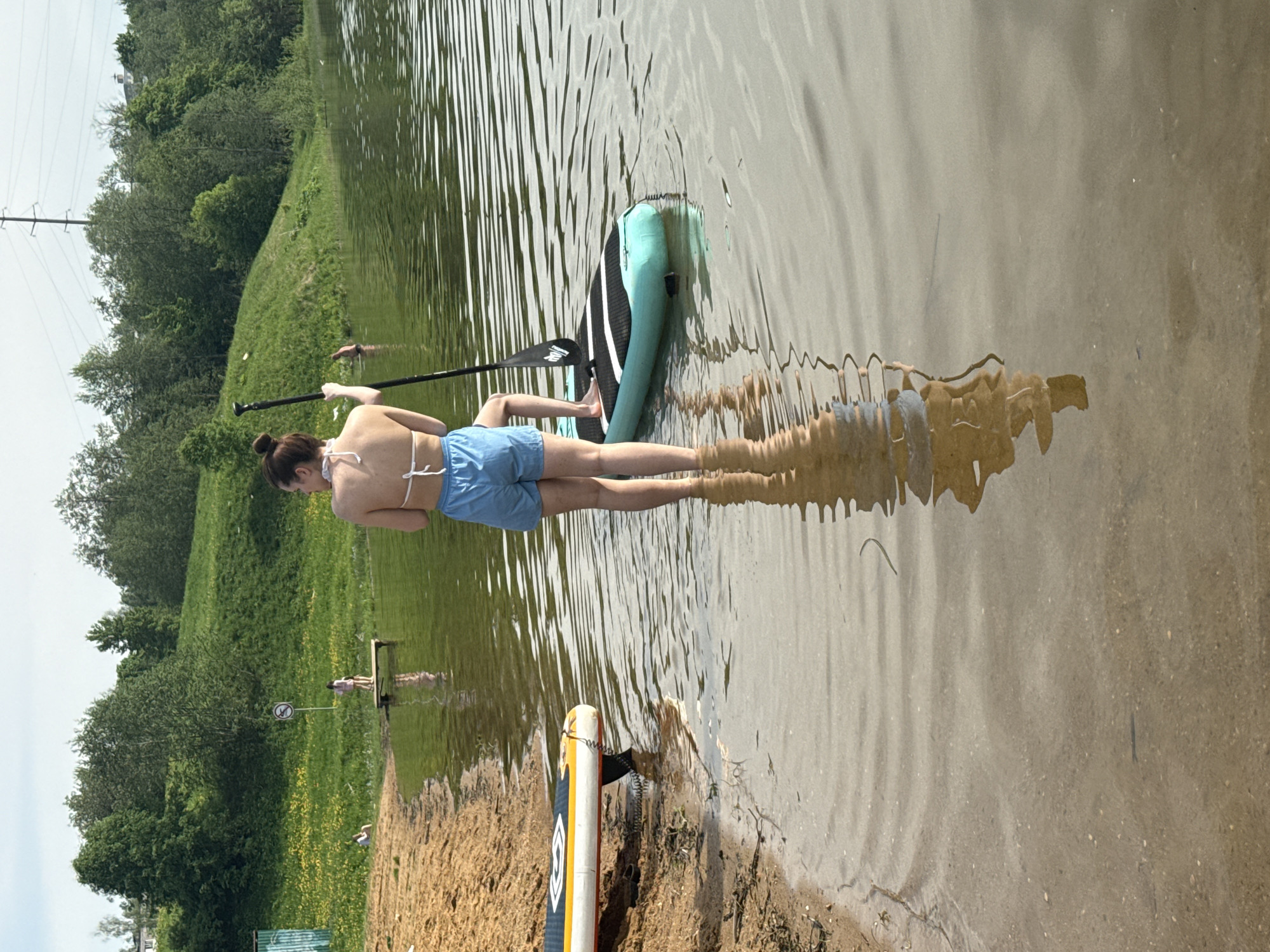 A woman in a white bikini top and blue shorts is lying prone on a paddleboard in a lake. She is holding a paddle. Another paddleboard rests on the sandy shore in the foreground. The lake is bordered by a grassy shoreline with trees in the background. The location is Novosinkovo, Russia.