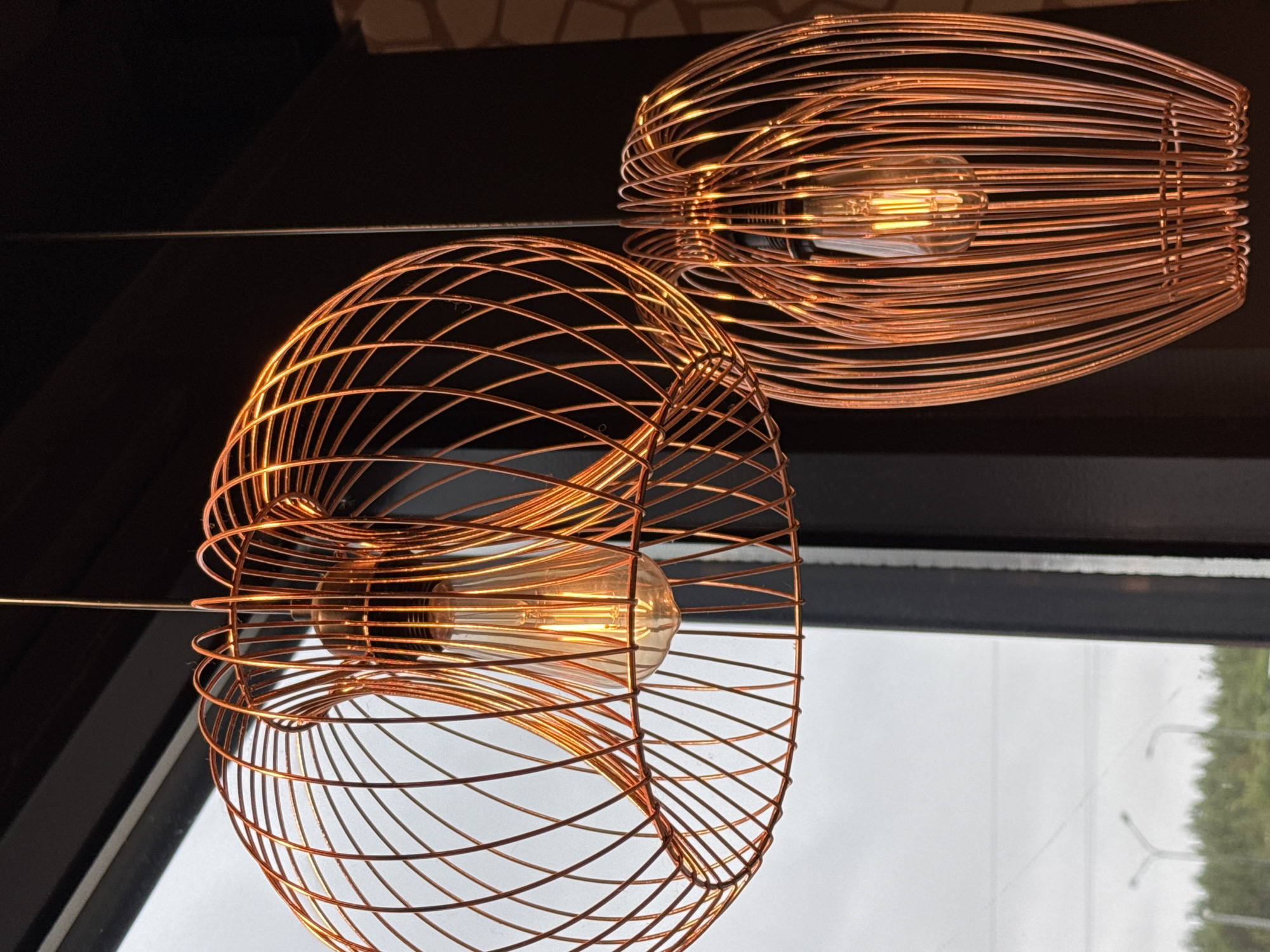The image shows two copper-colored wire cage pendant lights. Each encloses a warm-toned light bulb. The foreground light is spherical, while the background light is oblong. They are suspended against a dark surface, possibly a ceiling, with a window visible below. The view from the window is indistinct but shows a cloudy sky and foliage. There is no definitive indication this is Medovoye, Russia.