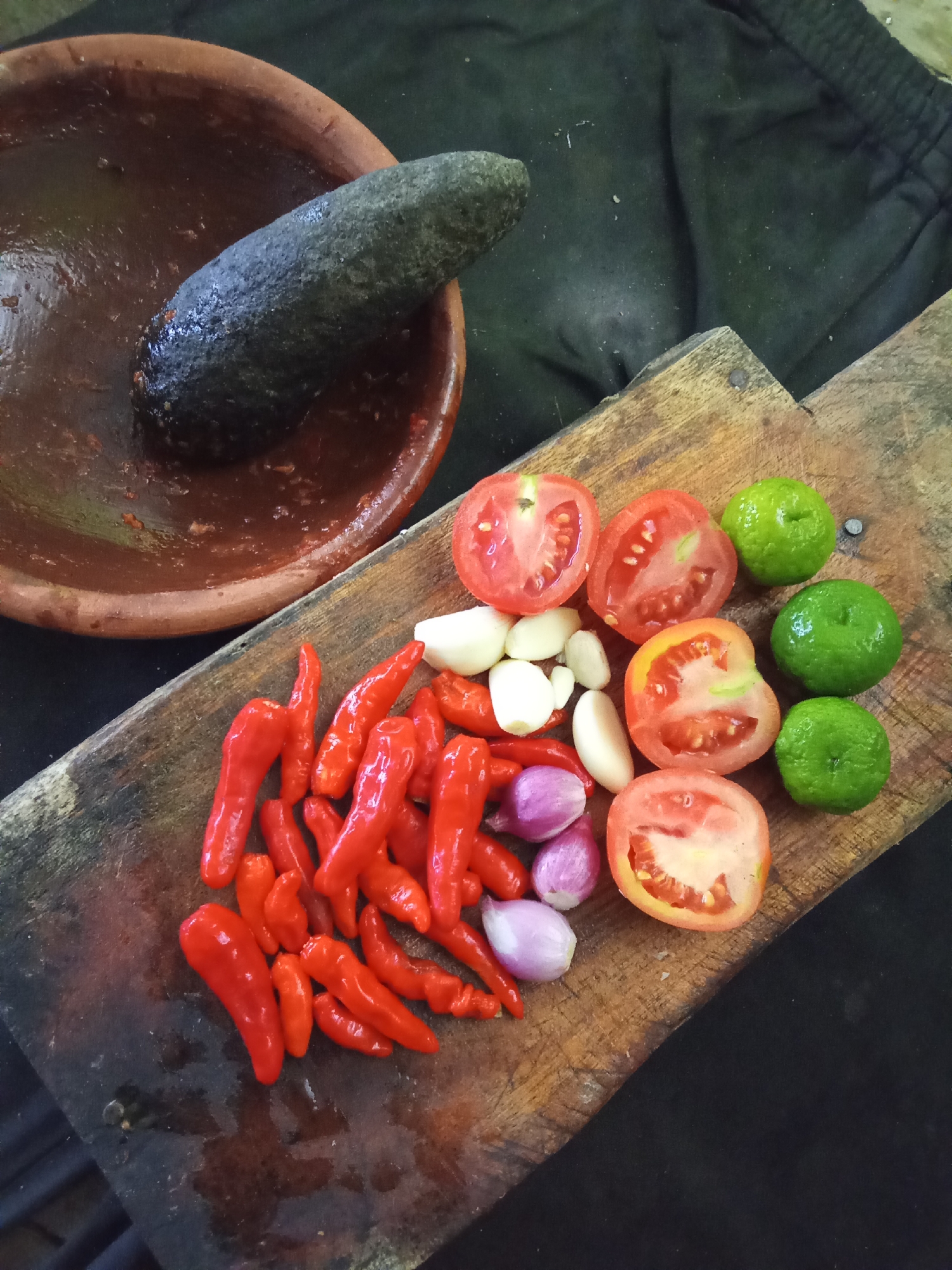 A mortar and pestle are shown next to a cutting board with several ingredients on it. The mortar is made of clay and contains some reddish residue. The pestle is made of dark stone. The cutting board is made of wood and appears weathered. On the cutting board, there are several red chilies, garlic cloves, shallots, sliced tomatoes, and small green limes. The ingredients are arranged in a somewhat scattered manner. The background is a dark, textured fabric. The image is shot from a high angle, looking down on the scene. The lighting is natural and appears to be coming from the side. There are no people or animals visible in the image. The scene appears to be a preparation area for cooking, likely in a home or small kitchen setting. The location is not specified in the image itself, and thus cannot be determined to be in Subang, Indonesia.