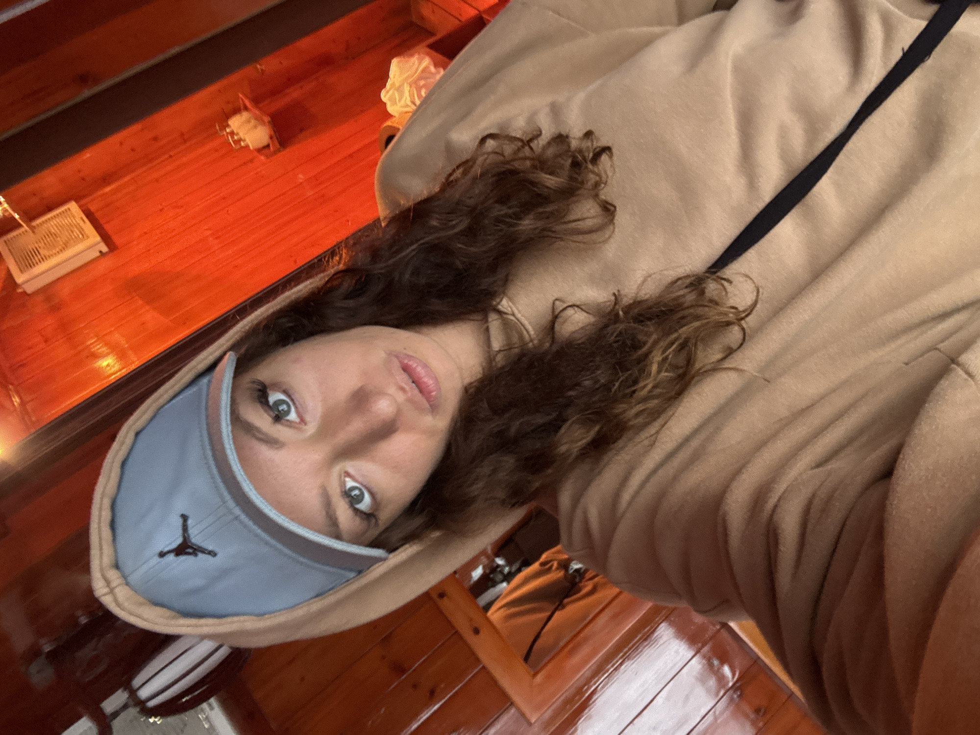A woman with curly brown hair and light eyes is wearing a blue baseball cap with a Jumpman logo and a tan hooded sweatshirt. The location appears to be inside a building with wooden paneling on the walls and ceiling. The light is somewhat dim and gives the wood a reddish hue. The woman is looking directly at the camera. The location is within Binh Lu, Vietnam.