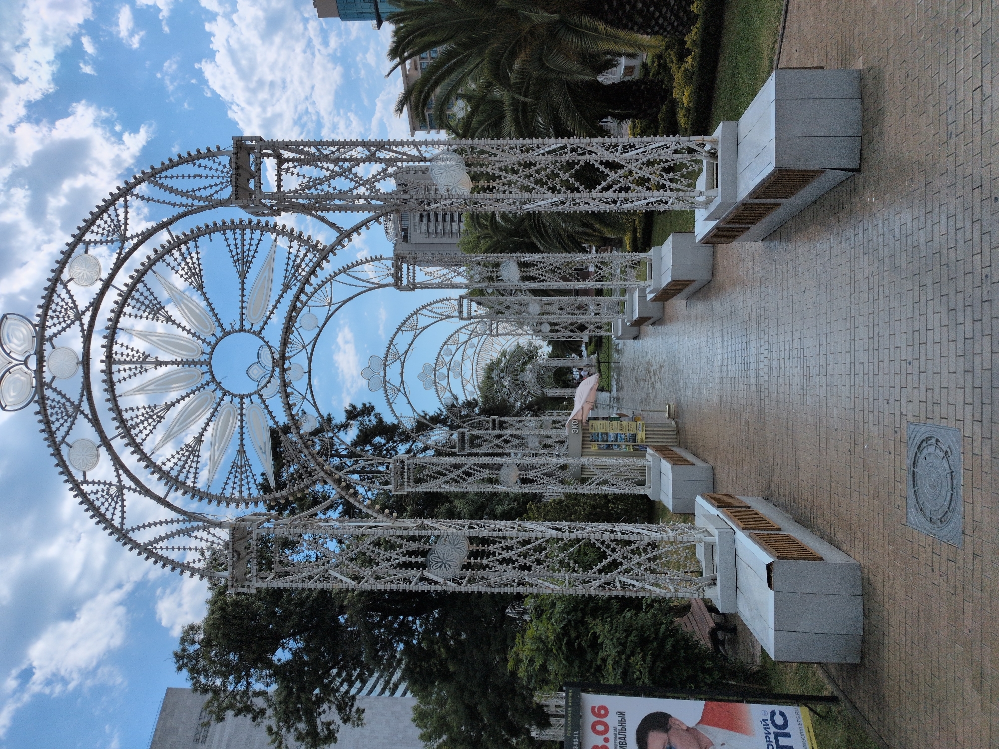 This is an outdoor, eye-level shot of a decorated walkway, likely in Sochi, Russia. The main subjects are ornate, white, metal archways that form a tunnel-like structure, adorned with intricate patterns and what appear to be decorative lights. The perspective is unusual, as the image is oriented vertically with the sky at the bottom and the ground at the top.
The walkway itself is paved with light-colored bricks. To the sides of the path are modern, minimalist benches made of concrete with wooden slats. Lush green trees and palm trees frame the scene, suggesting a warm climate. In the background, modern buildings are visible, hinting at an urban or resort setting. The sky is blue with scattered white clouds, indicating it's daytime, likely sunny.
There are no people prominently featured, though a small, light pink umbrella can be seen in the distance within the archway. A poster is visible at the bottom right corner with Russian text, including "3.06" and what appears to be "РИВАЛЬНЫЙ". Another part of the poster shows a portion of a man's face. The overall mood is serene and festive, with the decorative archways suggesting a special occasion or a well-maintained public space. The time of day appears to be afternoon given the light and shadows.