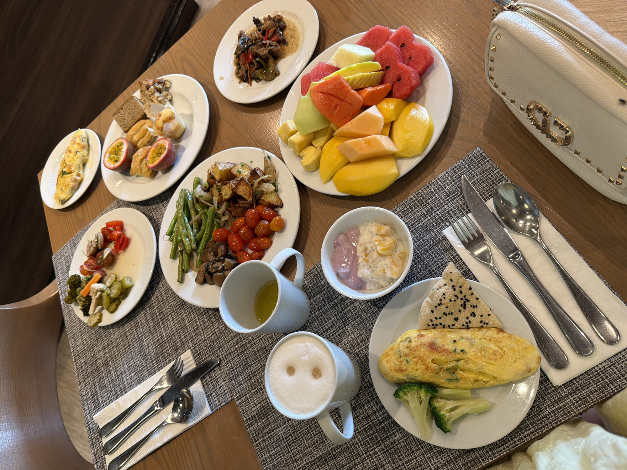 This image depicts a breakfast spread laid out on a wooden table. The setting appears to be a restaurant or hotel dining area in Bang Muang, Thailand, indicated by the specific style of wooden furniture and placemats.
In the foreground, a white plate holds an omelet, a side of broccoli florets, and a piece of crackers topped with black sesame seeds. Next to this plate are three pieces of silverware, a fork, a knife, and a spoon, neatly arranged on a white napkin. To the left, a white mug contains a frothy beverage, possibly coffee or a latte, with two small indentations on its surface. Beside it, another white mug holds a yellow liquid, likely juice or tea. A small white bowl contains a pink yogurt layered with what appears to be chia seeds and fruit.
Further back, a plate contains a colorful assortment of sliced fruits including mango, papaya, cantaloupe, pineapple, and watermelon, cut into heart shapes. Another white plate holds green beans, cherry tomatoes, and roasted potatoes. A plate in the upper left displays a medley of pickled vegetables and cherry tomatoes. In the upper right, a plate features what seems to be stir-fried beef with peppers and onions, accompanied by a slice of bread and half a passion fruit. An omelet is also visible on a plate in the upper middle of the image.
A light-colored clutch purse with gold studs and a prominent gold "G" emblem rests on the table to the right. The overall scene suggests a well-presented breakfast buffet. There are no people visible in the image.