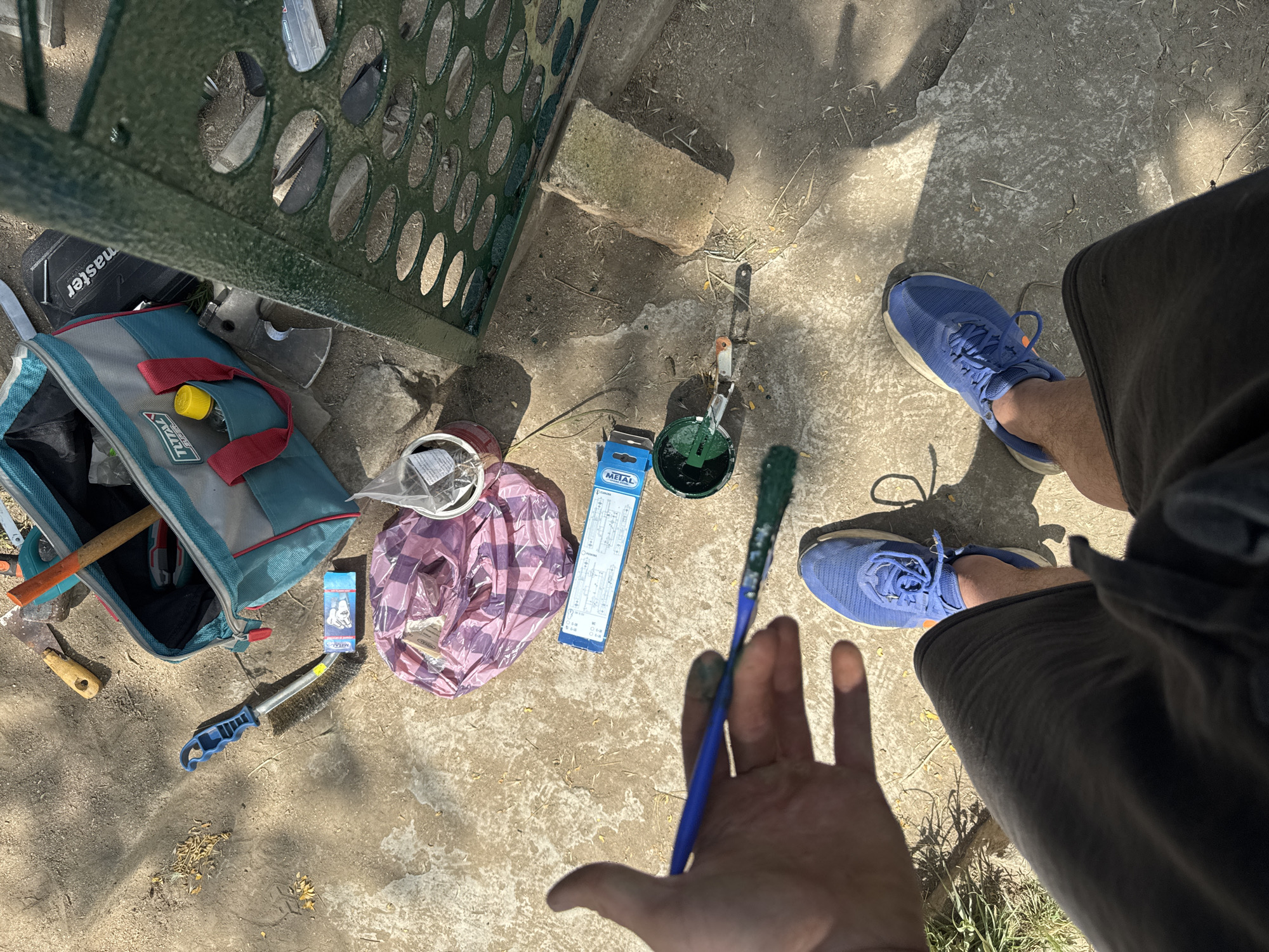 The image shows a top-down view of tools and supplies laid out on a concrete ground, likely outdoors given the shadows and sunlight. The scene appears to be set in Varna, Bulgaria.
In the foreground, a person's hand is visible, holding a paintbrush with green paint on its bristles. The hand is slightly smudged with paint, suggesting an ongoing activity. Next to the hand, a pair of blue athletic shoes are prominent. The person is wearing dark shorts.
Scattered around the ground are various items related to painting or maintenance. There is a teal and grey tote bag, possibly a tool bag, with a yellow-capped bottle inside. A few tools are visible, including what appears to be a wire brush with a blue handle, a small axe or hatchet, and a hammer with an orange handle. A roll of what looks like plastic sheeting or a bag of some sort, in a pink and purple plaid pattern, is also present. A small blue and white box with text is near the wire brush.
In the background, a green metal structure, possibly part of a fence or bench, is partially visible. A rectangular concrete block is also part of the scenery. The ground is concrete with scattered debris, such as dry leaves and twigs. The lighting suggests it is daytime, with strong sunlight casting distinct shadows. The overall impression is of a DIY project or outdoor repair work in progress.