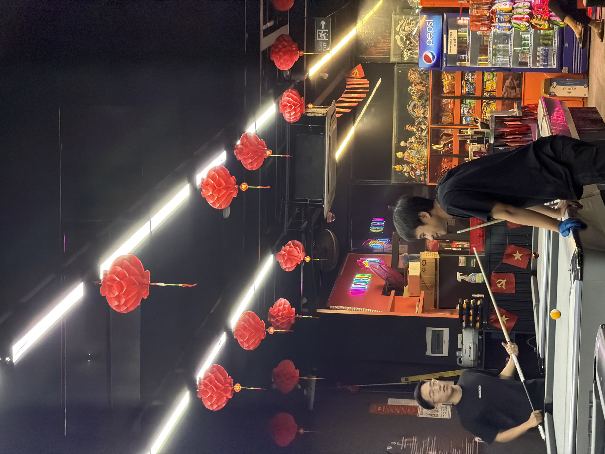 The image captures an indoor scene within what appears to be a billiards hall or entertainment venue, consistent with the provided location context of Đà Nẵng, Vietnam. The setting is dimly lit with overhead bright linear LED strip lights and numerous red, pleated paper lanterns hanging from the ceiling, adding a cultural or festive aesthetic.
Two young men are actively playing billiards. The man in the foreground, wearing a black t-shirt and glasses, is focused on lining up a shot on a pool table, holding a cue stick. A yellow pool ball is visible on the table. Further back, another man, also in a black t-shirt, is bent over a different pool table, seemingly engaged in a shot or observing the game. Both appear concentrated on their activity.
The background reveals several details characteristic of such a venue. On the right, shelves are stocked with various packaged snacks, and a Pepsi branded cooler filled with drinks is visible. Text on the cooler clearly reads "PEPSI" and "THÀNH TOÁN NƯỚC TẠI QUẦY" (Pay for water/drinks at the counter), confirming the Vietnamese language and context. Small red flags with a yellow star, some featuring a hammer and sickle emblem (flags of Vietnam and the Communist Party of Vietnam), are draped near one of the pool tables. Other elements include colorful neon signs in the distant background, a cleaning spray bottle on a shelf, and an "EXIT" sign partially visible. The overall atmosphere suggests an evening or night setting for recreational activities.