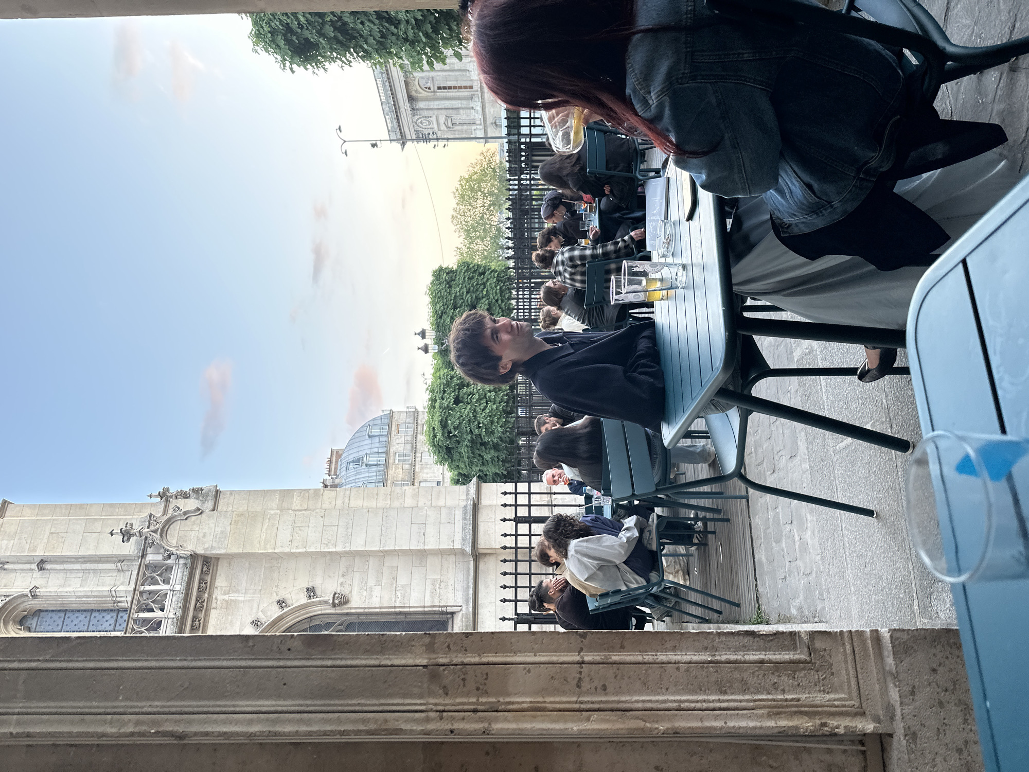 The image, oriented horizontally, captures an outdoor cafe scene in Paris, France, during what appears to be late afternoon or early evening. The sky is a pale blue with faint, scattered clouds, suggesting clear but possibly overcast weather.
In the foreground, a light blue outdoor table occupies the right side, partially obscured by an adjacent structure. A person with long reddish-brown hair, wearing a denim jacket over a light-colored dress or skirt, is seated at this table with their back to the viewer. They are holding a glass with a light-colored beverage, possibly beer or cider.
Further into the mid-ground, several other individuals are seated at similar light blue tables with dark chairs, arranged in a row along a stone pathway or patio. They are engaged in conversation, suggesting a social gathering. One man with dark hair, wearing a dark shirt, is prominently featured, looking towards the left side of the frame. Other patrons are visible further down the row, some with drinks on their tables.
The background features a blend of historical architecture and greenery. To the far left, a grand stone building with ornate Gothic or Neoclassical elements, including intricate carvings, decorative windows, and arched structures, rises prominently. Beyond the cafe seating, a dark metal fence separates the seating area from a line of green trees. Further back, another building with a distinctive domed roof can be seen, along with a street lamp. The overall atmosphere is relaxed and social, characteristic of an urban outdoor dining experience. No discernible text is visible within the frame.