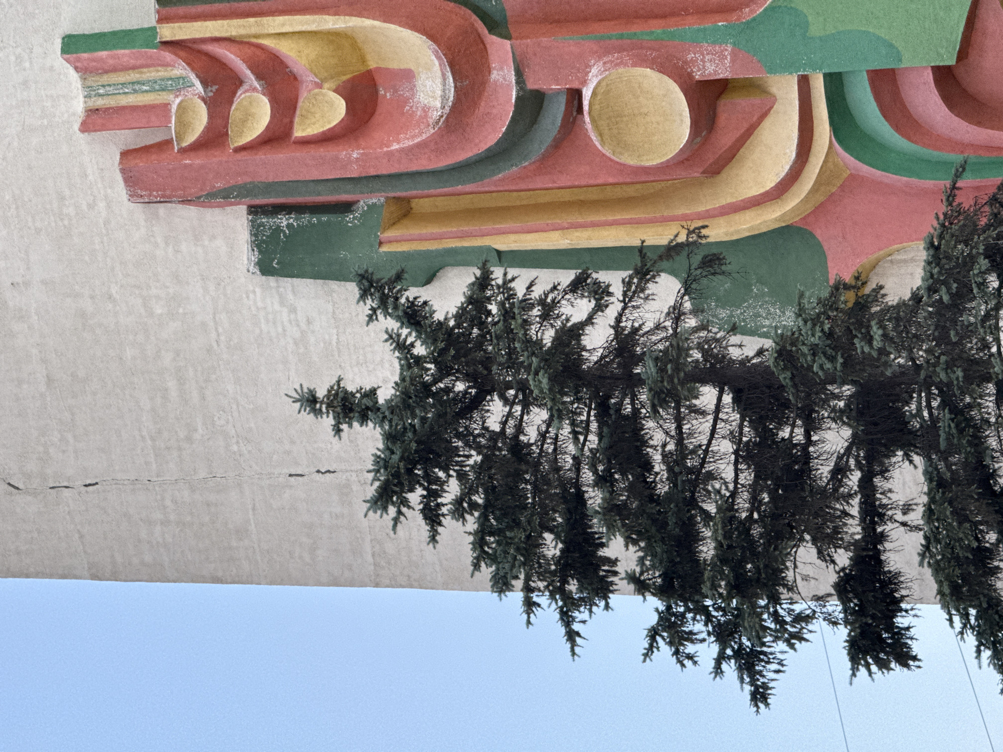 This media file depicts a close-up, low-angle view of an outdoor architectural feature and overhead evergreen branches against a bright blue sky. The architectural element is a textured surface with a distinct, layered design in shades of red, yellow, and green. The shapes are curved and geometric, with rounded indentations and raised sections, suggesting a decorative mural or facade. The surface appears weathered, with visible specks of white possibly indicating paint chips or dust. The branches of what appear to be pine or fir trees hang down into the frame, obscuring parts of the architecture and the sky. The overall impression is one of an abstract art installation or a unique building detail situated in a natural environment. The lighting suggests it is daytime, likely under clear skies. The rotation of the image suggests it may have been taken from below, looking upwards. No people, text, or specific events are discernible. The location context of Kokshetau, Kazakhstan, offers a potential setting for such a structure, possibly a public monument or a building with distinctive ornamentation.