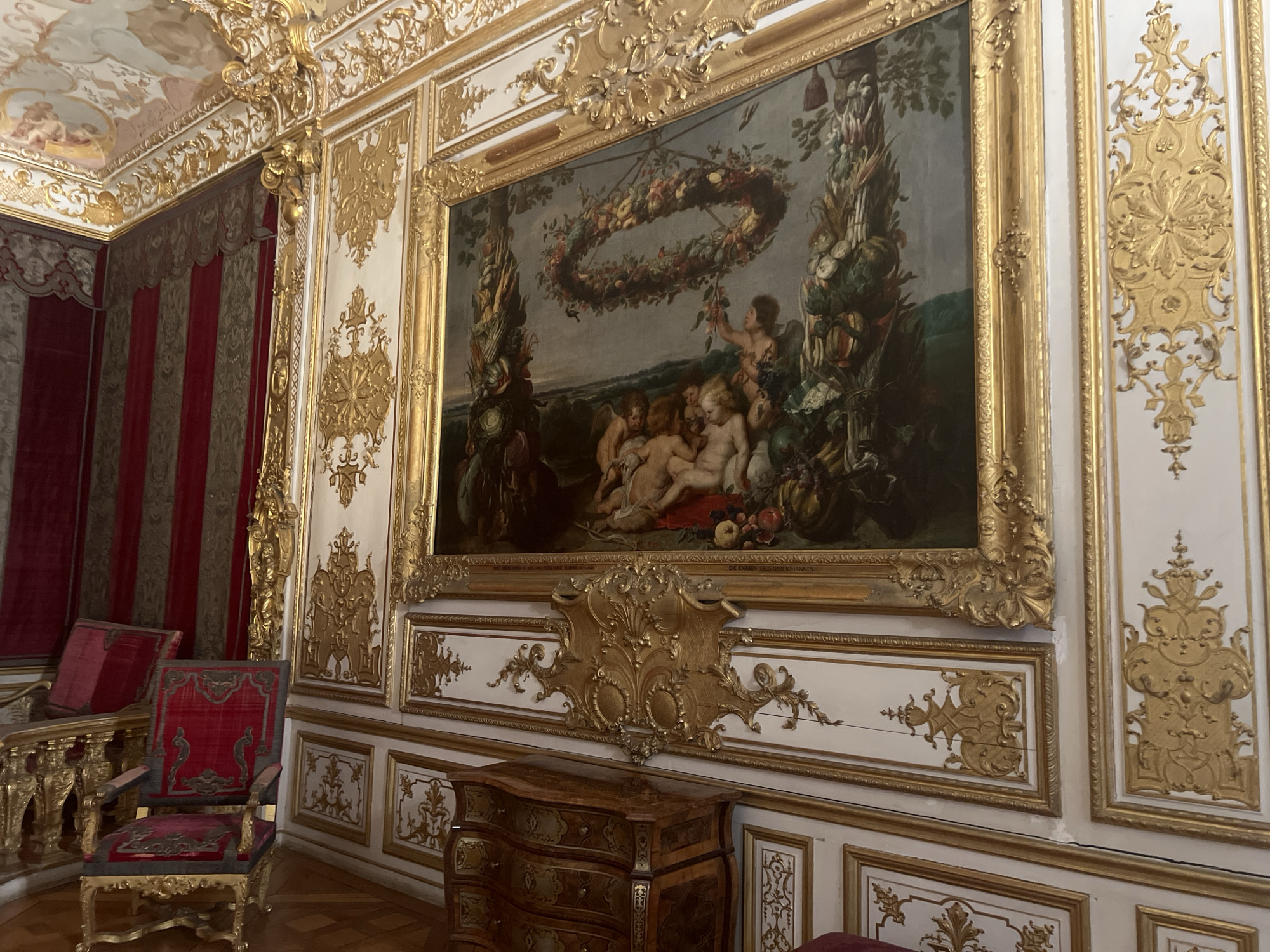 The image depicts an opulent interior within the New Schleißheim Palace in Oberschleißheim, Germany. The scene is dominated by an ornate, gilded rococo-style room. A large, framed painting hangs prominently on the wall, depicting cherubs adorning a garland of fruit and vegetables. The painting is surrounded by elaborate gold trim and ornate decorative panels. To the left of the painting, red velvet draperies with vertical stripes are visible, partially obscuring another section of the room. Two red upholstered chairs with gilded frames are positioned in the foreground on the left. On the right side of the painting, more gilded decorative panels are mounted on the white walls. Below the painting, a small, ornate wooden chest of drawers with intricate marquetry is visible. The floor is made of polished wood planks. The ceiling, visible in the upper left corner, features painted frescoes with putti and is also adorned with gilded decorations. The overall impression is one of extreme wealth and elaborate artistry characteristic of Baroque or Rococo palace interiors.